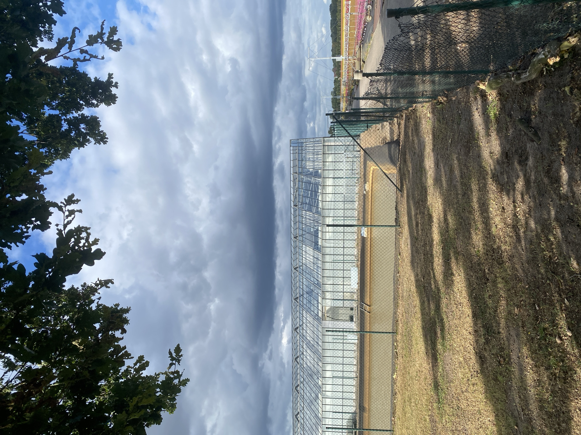 The image shows a long, modern greenhouse structure with a glass roof and white walls, located in Schwarme, Germany. The greenhouse is surrounded by a chain-link fence and a muddy area with dry grass. In the background, there is a walkway or road with some equipment and structures, including a suspension bridge-like structure and what appears to be colorful carnival rides or stalls. Overcast skies with dramatic clouds loom above the scene, contrasting with patches of blue sky. Tree branches with green leaves hang from the top left corner of the frame. A ladder is visible propped against the wall of the greenhouse inside the fenced area.