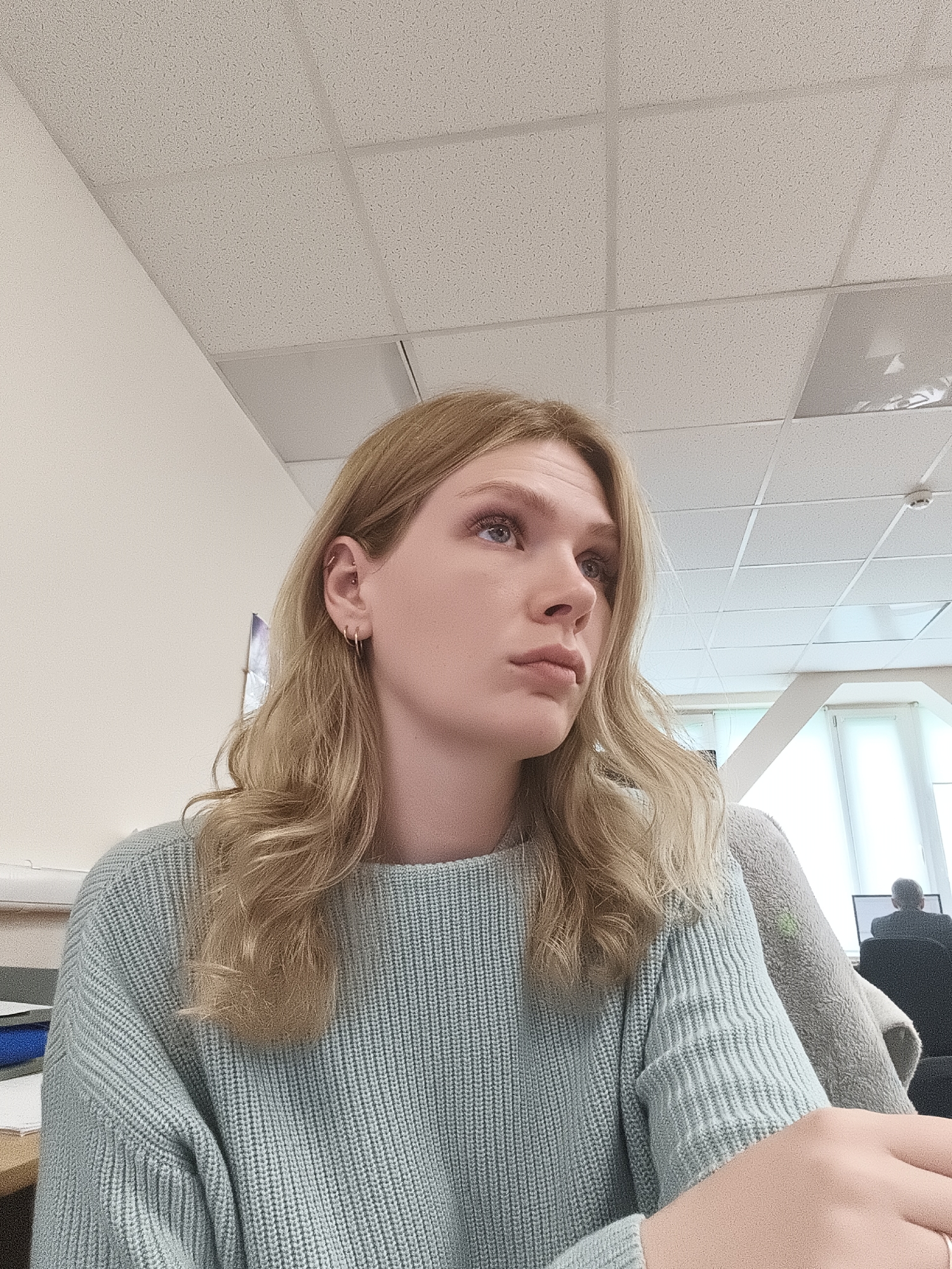 A fair-skinned woman with long, wavy blonde hair sits at a desk. She is wearing a light blue, knit sweater. She has multiple piercings in her left ear. Her gaze is directed upwards and to the right, and her lips are pursed in a thoughtful expression. The environment appears to be an office setting, with a tiled ceiling, fluorescent lighting, and a white wall in the background. Another individual can be seen in the background, seated at a computer. The location is Unknown, Unknown.
