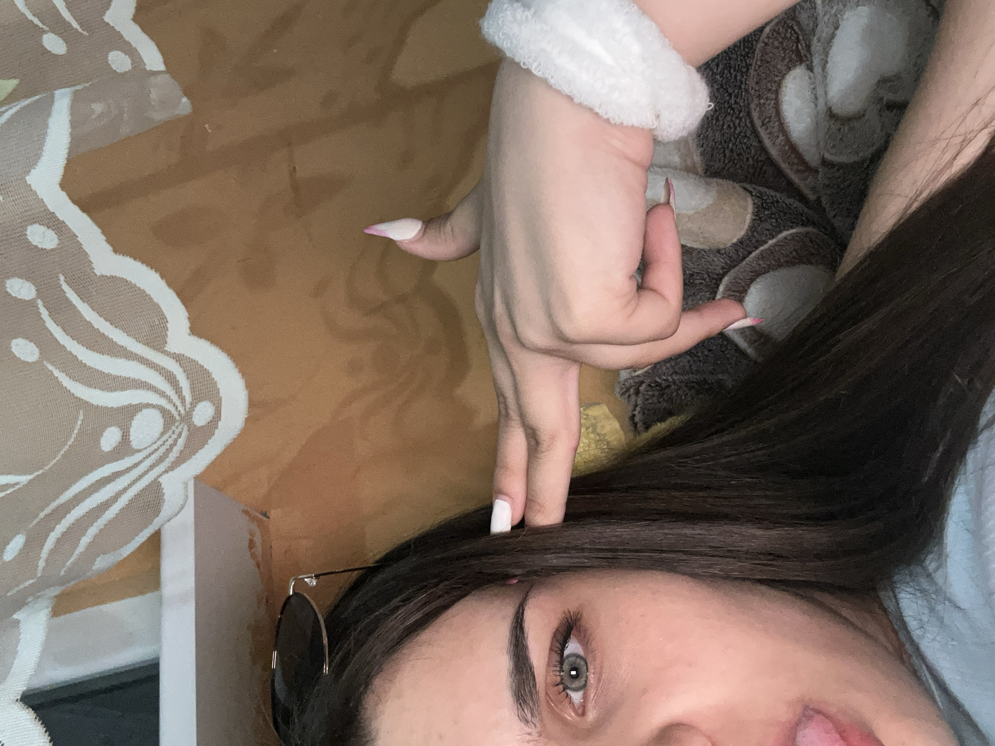 The image shows a close-up of a young woman's face and hand. She has dark hair, light skin, and grey eyes. Her tongue is sticking out, and her right hand is raised to her head, with her index finger and thumb forming a gun shape. She is wearing a white furry bracelet on her left wrist. The background appears to be a bed with a patterned blanket and a piece of lace fabric draped over it. There are no identifiable landmarks or features that would indicate a specific location in Shumen, Bulgaria.