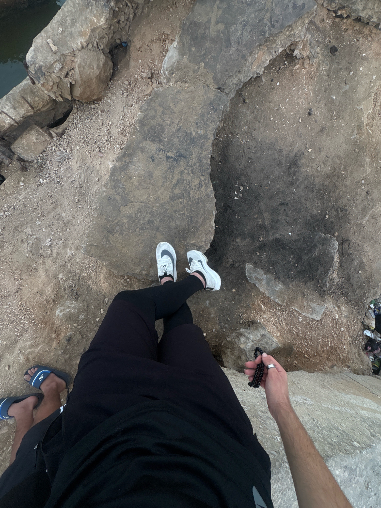 A first-person perspective shot shows a person sitting on a rocky ledge, facing away from the camera and looking down at the ground. They are wearing black pants, white sneakers, and holding a string of black beads in their right hand. To the left of the frame, another person's legs are visible, wearing blue slides and black shorts. The ground is covered in sand, gravel, and large rocks. In the background, there is a body of water and some debris. The overall environment appears to be a coastal area.