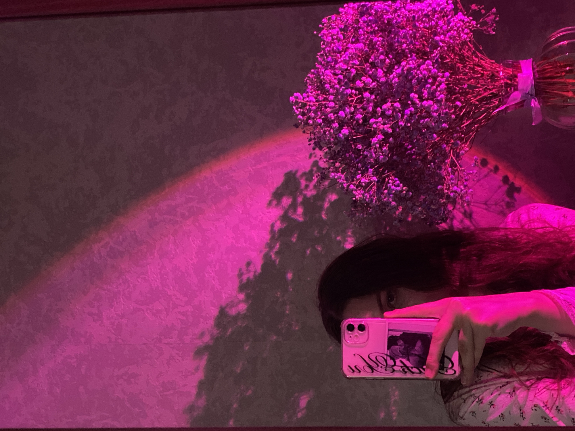 A person is holding a pink smartphone, seemingly taking a picture of themselves in a mirror. The person has dark hair and is wearing a patterned shirt. A bouquet of purple flowers in a glass vase is in the upper right corner. A pink light is cast on the wall behind the person and flowers. The location of the image is unknown and cannot be determined based on the picture alone. The city of Izhevsk, Russia, also cannot be verified.