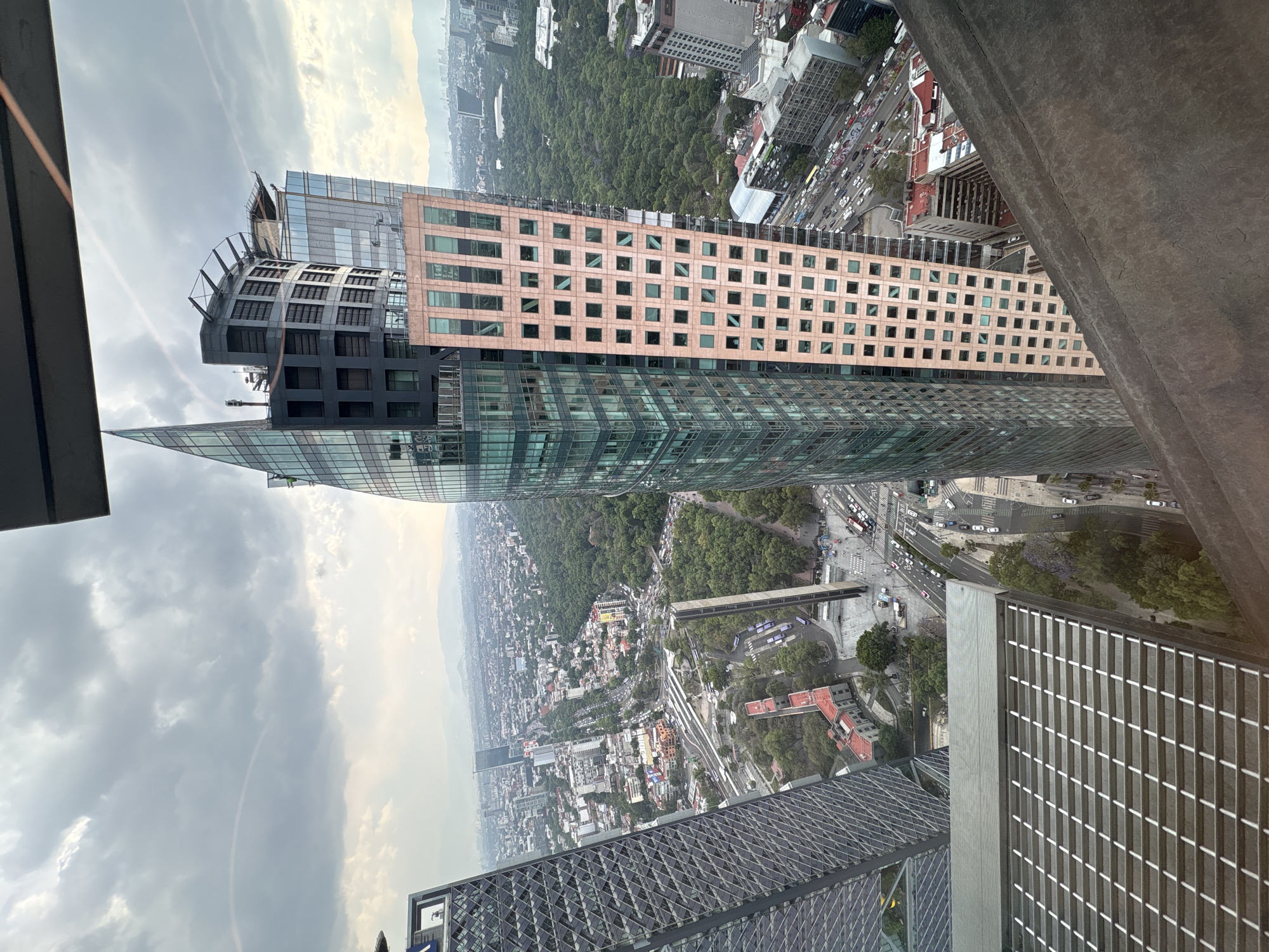 The image depicts a high-angle view of Mexico City, Mexico. The foreground is dominated by the edge of a modern building, possibly the viewpoint from which the photo was taken. Another tall building, characterized by a combination of glass and light pink facade sections with regularly spaced windows, is prominent in the middle ground. Beyond the buildings lies a sprawling urban landscape interspersed with patches of dense greenery, likely parks or forested areas. A major road with vehicular traffic can be seen snaking through the city. The sky above is overcast with grey and white clouds.