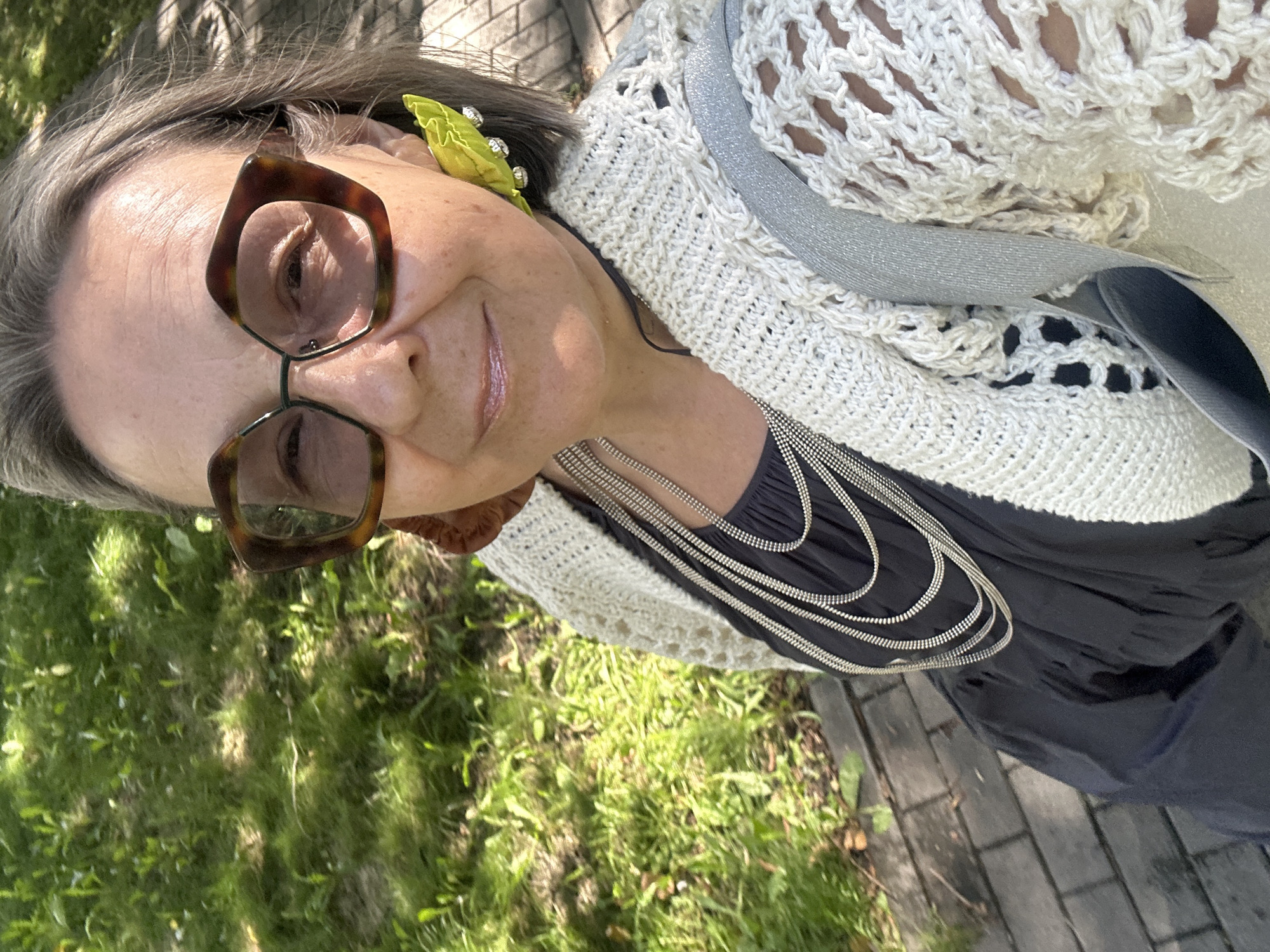 A medium shot photo shows a woman with gray hair, wearing oversized, tortoise-shell glasses, and large, lime green leaf-shaped earrings. She is dressed in a dark dress layered with a cream-colored, crocheted cardigan. Several silver necklaces are layered over her dress. The woman is smiling and looking at the camera. She appears to be outdoors, with blurred green grass and a brick pathway visible in the background. The lighting suggests it is daytime, with sunlight casting shadows on the grass.