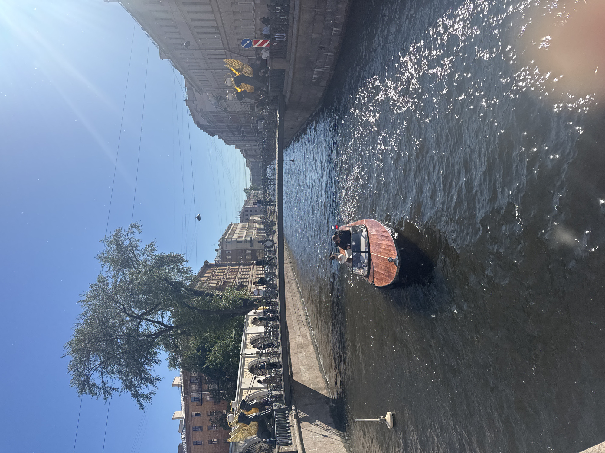 The image depicts a canal in Saint Petersburg, Russia, likely the Griboyedov Canal given the architectural style and the presence of the Lion Bridge (also known as Bank Bridge) in the background, identified by the griffins on either side of its approaches. A narrow wooden motorboat, with several passengers, is navigating the canal. People are visible on a pedestrian bridge spanning the canal and on the canal's embankment. The buildings lining the canal are typical of Saint Petersburg's historic architecture, with multiple stories and ornate facades. The sky is clear and blue, indicating a sunny day, with the sun casting reflections on the water's surface. The overall scene is one of urban activity and sightseeing on a bright day.