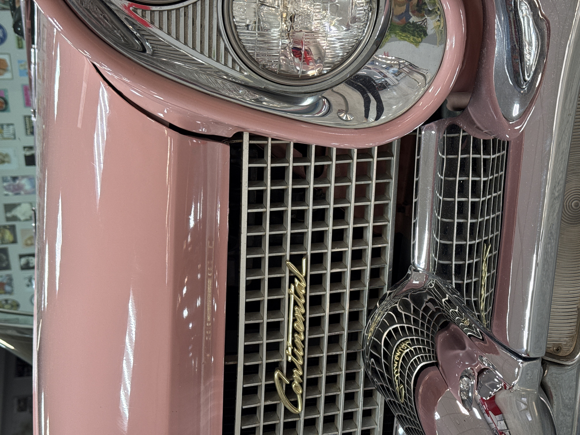 This close-up image showcases the front right quarter of a classic car, featuring a striking glossy pink (or salmon) paint finish. The car is adorned with chrome accents and a distinctive grille.
The main subjects are the car's components: a circular headlight with a reflective lens and chrome bezel, a square-patterned grille, and highly polished chrome trim elements. The pink bodywork reflects vertical streaks of light, indicating an indoor setting with overhead illumination. Reflections are prominent throughout the image; the chrome bezel of the headlight shows a distorted view of a person's legs and torso, while other chrome surfaces capture various environmental elements.
A key detail is the car's emblem, a gold script reading "Continental," positioned vertically on the grille. The grille itself features a regular, intricate grid pattern. The vehicle appears to be stationary, likely on display. The background, subtly reflected in the glossy surfaces and visible to the far left, suggests an indoor location such as a museum, showroom, or collection, with what looks like framed pictures or display items on a wall. There are no explicit people visible, but reflections suggest an observer or photographer. The image conveys a sense of vintage luxury and careful preservation, set in Sochi, Russia.