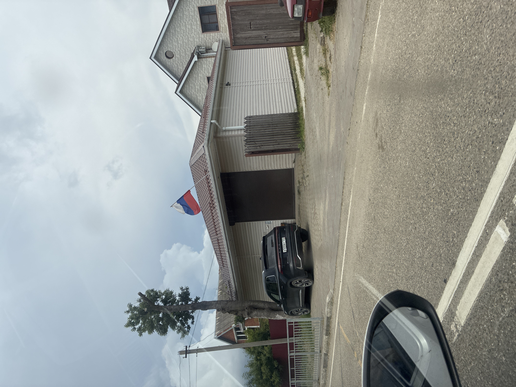 The image depicts a street scene in Arkhipo-Osipovka, Russia. A black SUV is parked on a paved area in front of a building with a light-colored vinyl siding and a dark roll-up door. A Russian flag flies from the roof of the building. To the left of the SUV, a tall, slender tree with a pruned top grows at an angle. Behind the building, a larger, multi-story house with light stone siding is visible. A red wooden fence runs along the property line on the left, and a portion of a red vehicle is visible on the right side of the image. The foreground shows a street with asphalt paving and white road markings, as seen through a car's side mirror. The sky is cloudy.