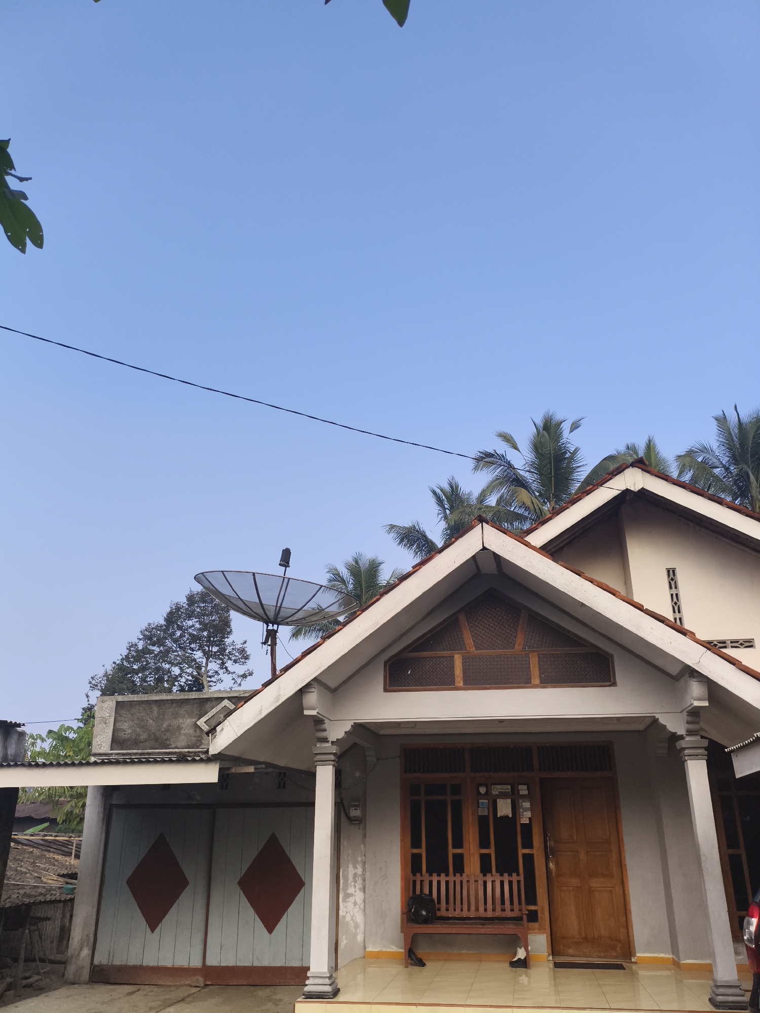 In the city of Kembaran, Indonesia, a house with a light gray facade is pictured under a clear blue sky. The house features a prominent pitched roof covered in red tiles and a triangular gable with wooden latticework. A satellite dish is mounted on a pole to the left of the house, and palm trees are visible in the background. The front of the house has a wooden door and a window with a wooden bench placed in front of it. To the left of the house, a set of light blue double doors with red diamond shapes is visible. A dark-colored bag rests on the bench, and two pairs of shoes are on the porch. The scene is serene, with no people or animals actively engaged in any activity.