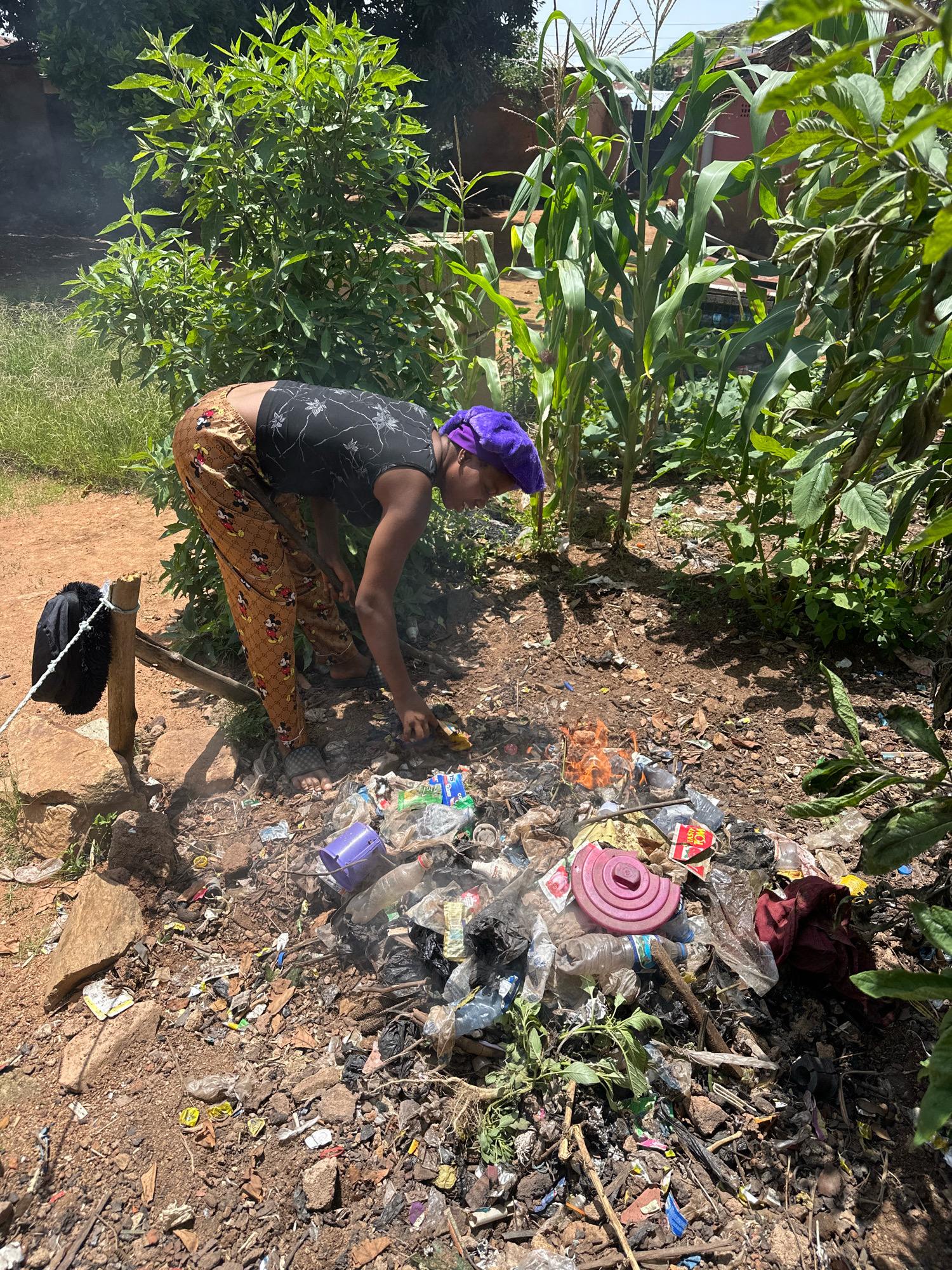 In Gura Top, Nigeria, a person with a purple headscarf and patterned pants is tending to a small fire where plastic waste is burning. The fire is surrounded by a large pile of trash, including plastic bottles, wrappers, and other debris. In the background, lush green vegetation, including corn stalks and leafy plants, thrives. A black bag hangs from a wooden post on the left side of the frame. The ground is a mixture of dirt and rocks, indicating an outdoor, possibly rural, setting. The scene appears to be an uncontrolled waste disposal site, common in many parts of the world.