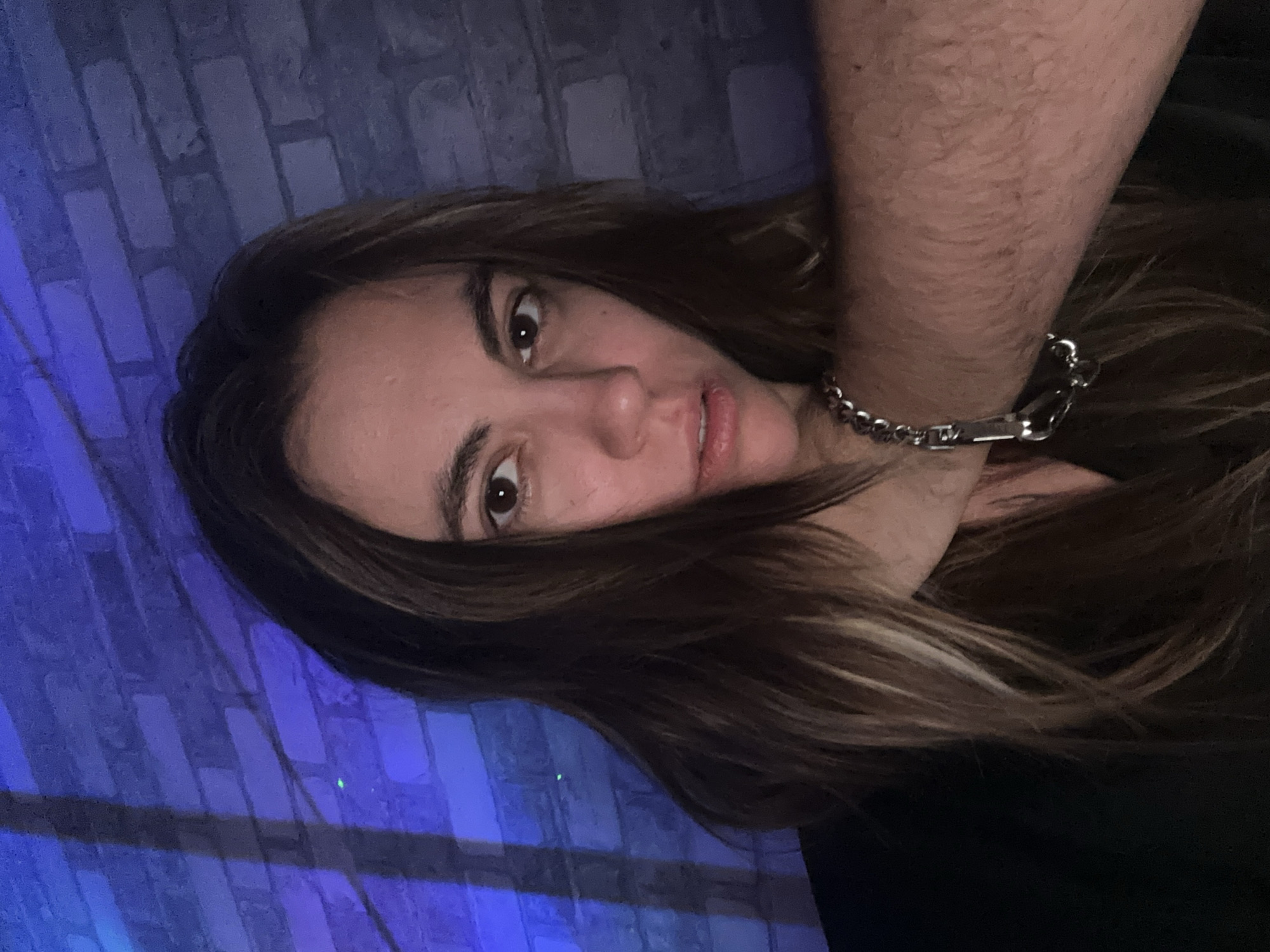 A woman with long, dark brown hair is shown from the chest up, facing the camera. Her eyes are looking directly forward. A tattooed arm with a silver bracelet is wrapped around her neck, and it is positioned across her throat. She is wearing a black garment. The background consists of a brick wall lit with blue and purple lights, casting shadows on the wall. The image appears to be a selfie taken in a dimly lit room. There is no indication of the location being Moscow, Russia.