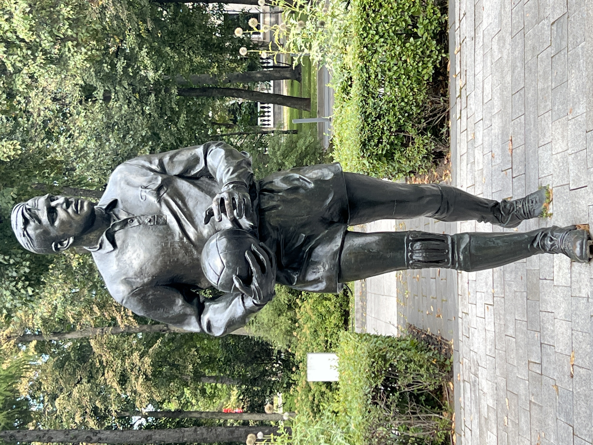 A full-body bronze statue of a male soccer player stands in a park. The statue is positioned in the center of the frame, facing forward and slightly to the right, holding a soccer ball in his hands. He is wearing a collared shirt, shorts, knee-high socks, and cleats. The background consists of lush green trees and manicured hedges, with a paved walkway in the foreground. The lighting suggests it is daytime.