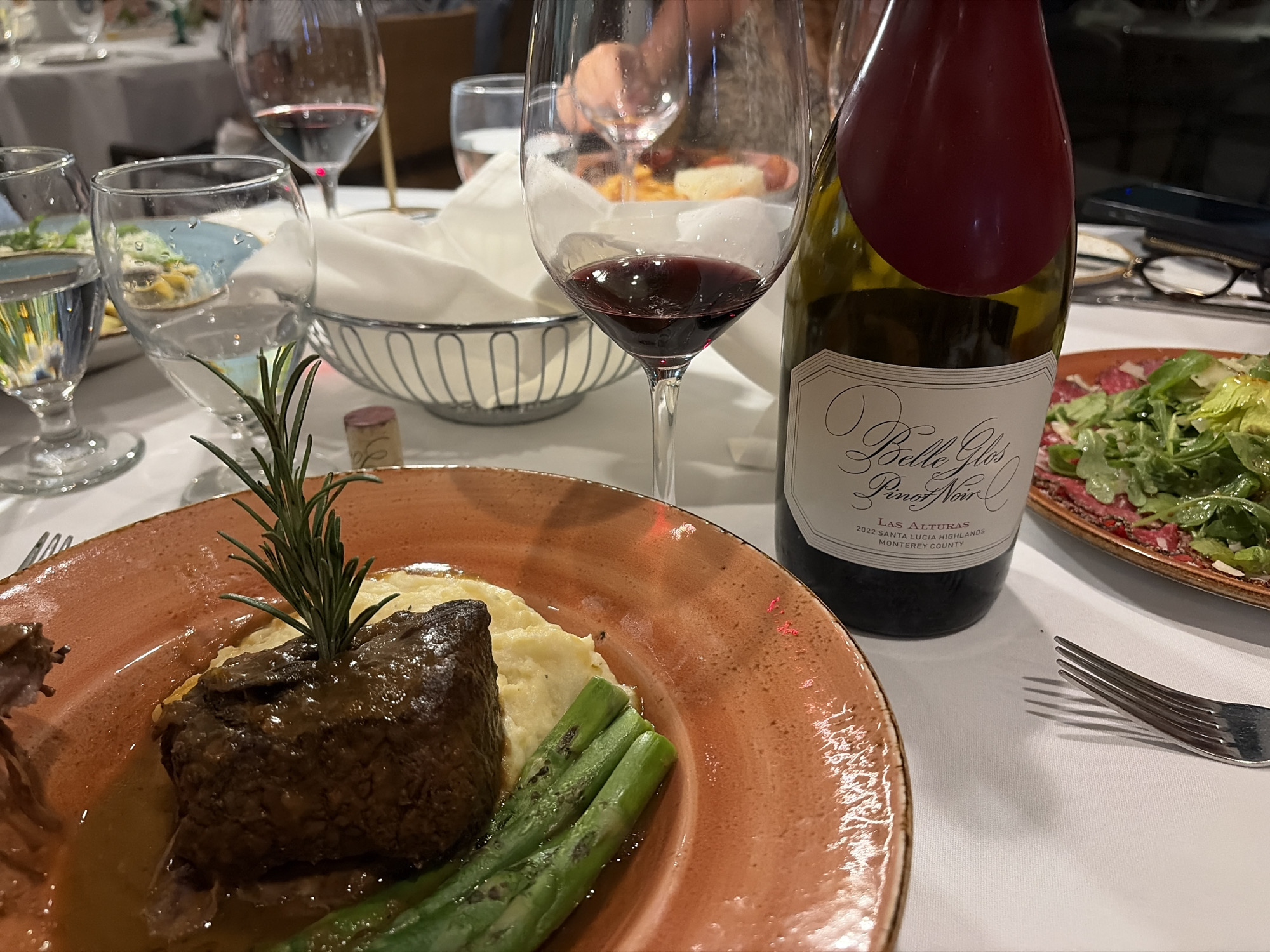 The image depicts a table setting in a restaurant, likely indoors, in an unknown city, United States of America. The scene is centered on a partially eaten meal: braised meat atop a bed of mashed potatoes, accompanied by asparagus, served on an orange-rimmed plate. A sprig of rosemary garnishes the meat. A bottle of "Belle Glos Pinot Noir Las Alturas 2022 Santa Lucia Highlands Monterey County" stands next to the plate, with a wine glass filled with red wine positioned in front of it. To the right of the wine bottle, another plate holds a salad with greens and a red topping. A fork is placed near the salad plate.
Other elements on the table include:
- a water glass with liquid
- a breadbasket with a white cloth napkin
- other wine glasses
- glasses
- other plates with food, only partially visible.
The lighting suggests it may be evening. The image does not reveal any information about the city or the restaurant, let alone Key Biscayne, United States of America, which are not verifiable details. There are no people visible.