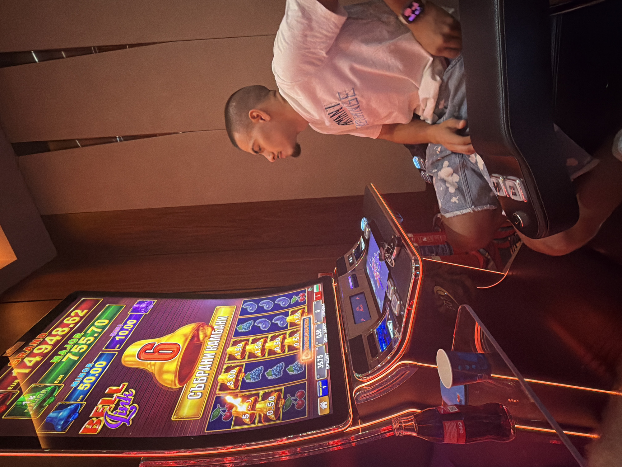 A person, likely male, is sitting in a casino playing a slot machine. The person is wearing a white t-shirt and blue shorts with a floral pattern. They are looking down at the slot machine, focused on the game. The slot machine has a colorful display showing various symbols, including bells, grapes, and cherries, and text in what appears to be Cyrillic script and English. The machine displays jackpot amounts like "GRAND 14948.62", "MAJOR 755.70", and "MINOR 50.00". There are also cups and a bottle of Coca-Cola on a surface near the machine. The background suggests an indoor setting, possibly a casino, with warm lighting and wooden paneling. The overall atmosphere seems relaxed, with the person engrossed in the activity. The visible text and potential Cyrillic script suggest the location is likely in Bulgaria, specifically Ruse, as indicated by the provided context. The lighting suggests it could be evening or night, a common time for casino activity.