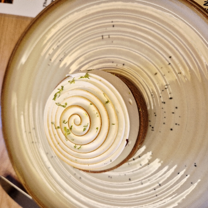 A close-up, high-angle photograph captures a single dessert presented on a ceramic plate. The dessert is a circular tart, likely a lemon meringue variety, with a visible light brown crust forming its base. A substantial layer of white meringue is meticulously piped on top in a continuous spiral pattern, originating from the center and expanding outwards. The outer edges and peaks of the meringue spiral exhibit light browning or toasting. Small, bright green zest fragments, consistent with lime or lemon, are scattered sparsely over the meringue's surface.
The dessert is centered on a round, beige-toned ceramic plate featuring a textured surface with concentric, raised ridges that create a ripple effect extending towards its darker brown rim. Irregularly shaped, small dark specks are scattered on the plate's surface, particularly in the lower left quadrant.
In the upper left portion of the image, a segment of a silver-colored cutlery handle is visible alongside the plate. The background in the upper left suggests a blurred light-brown wooden table surface. In the lower right corner, a portion of a blurred white object with faint black text is discernible, consistent with a menu or table information card. No individuals are present in the frame. The scene indicates a dining environment within a restaurant or patisserie.
**Location in the city of Strasbourg, France:** The image depicts a dessert served in a dining establishment located within the city of Strasbourg, France. The precise name or street address of the establishment is not discernible from the visual information provided.