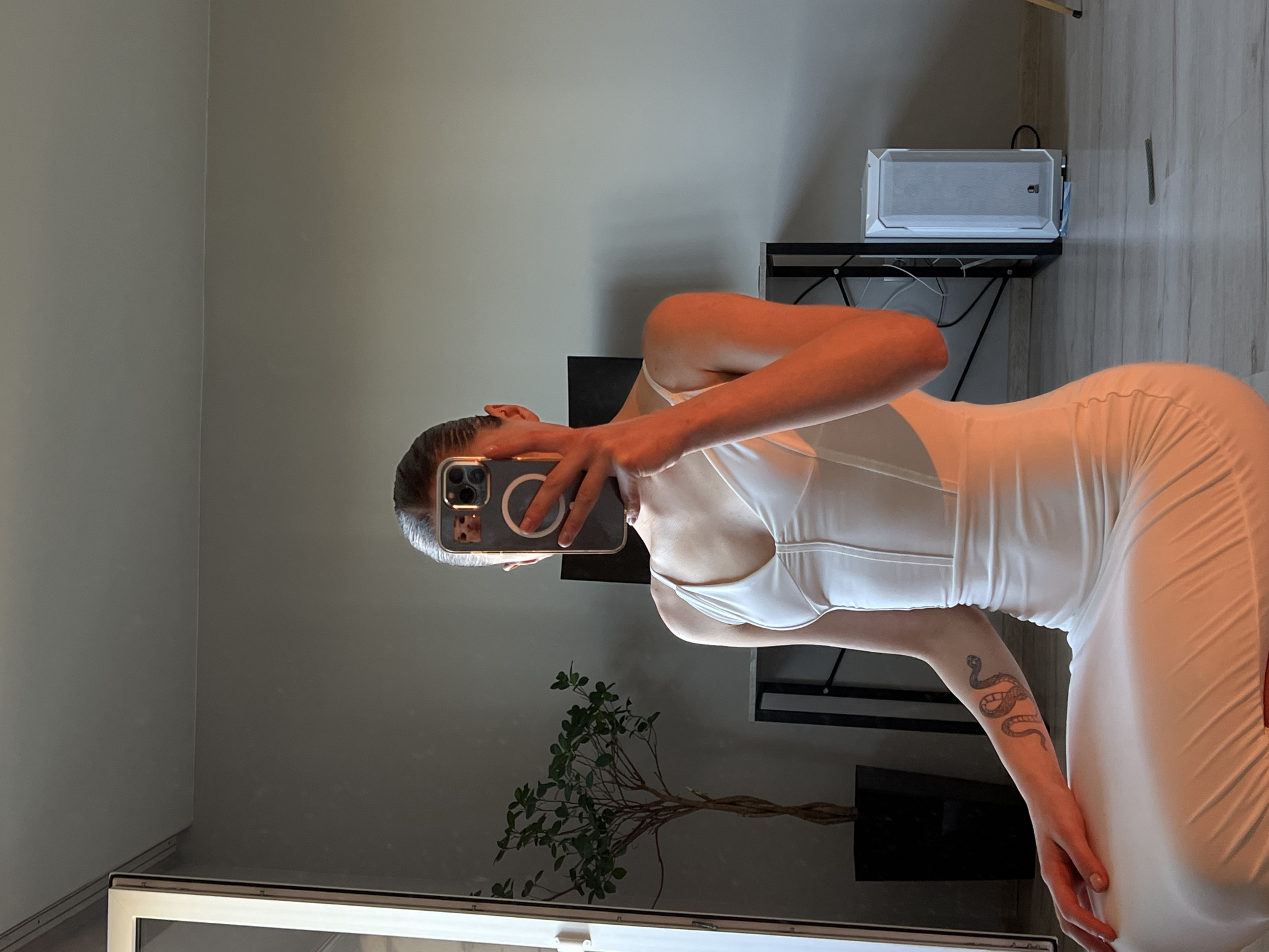 A young woman, seen from the chest down, sits on the floor in a room. She is holding a smartphone to her face, obscuring her features, and appears to be taking a selfie in a mirror. She is wearing a white, form-fitting dress with a plunging neckline and thin straps. A snake tattoo is visible on her left forearm. In the background, to her left, is a large mirror reflecting a potted plant and part of the room. To her right is a desk with a computer tower and cables. The room has light gray walls and a light-colored floor. The lighting suggests it is daytime, with warm sunlight casting orange hues on the woman's right side. The image does not provide information about the city of Ternopil, Ukraine.