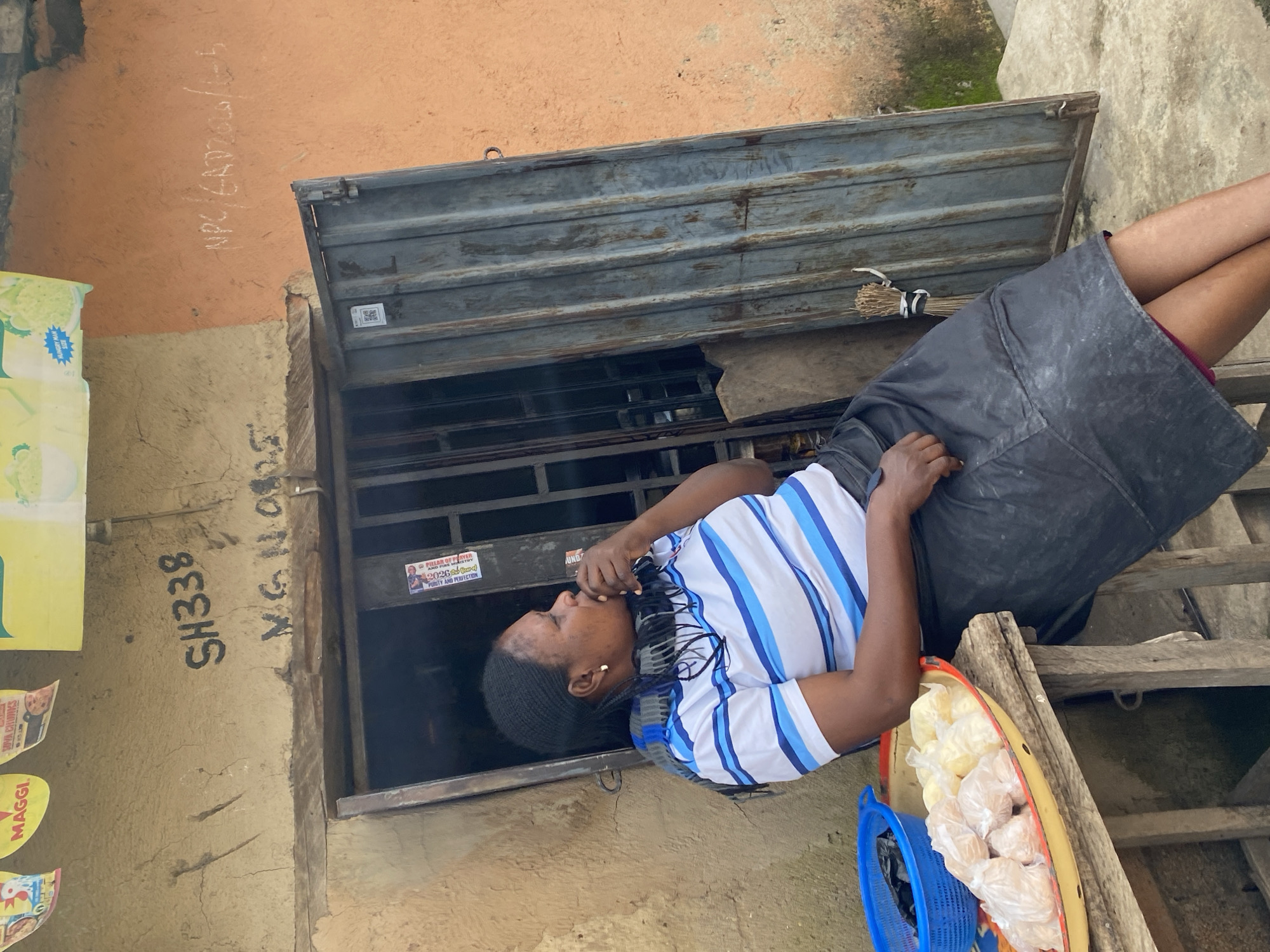A woman is seen sitting next to a storefront in Kabong, Nigeria. She is wearing a blue and white striped shirt and a black skirt. In front of her, on a wooden table, is a tray filled with small, individually wrapped food items, and a blue basket containing a black plastic bag. The storefront has a corrugated metal door, part of which is open. Above the door, graffiti is visible on the wall, including the markings "SH338" and "M.G.N 08." Another inscription, "NPC/ENP2020/6-5," is written in chalk on the orange wall to the right. The overall environment appears to be an outdoor market setting.
