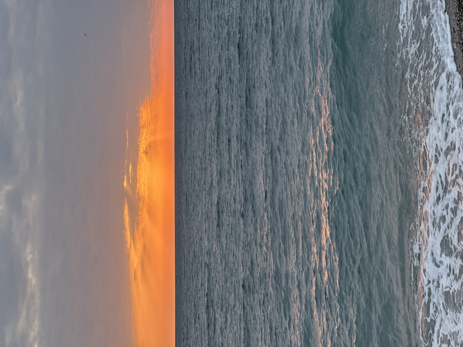The image displays a seascape at sunset or sunrise from the coast of Šušanj, Montenegro. The sky occupies the upper third, featuring a band of intense orange and red hues along the horizon, indicating the sun's position, with crepuscular rays emanating upwards through scattered clouds. Above this, the sky transitions to overcast grey clouds, and a single, small, dark, indistinct flying object is visible in the upper right quadrant. The middle section is dominated by the sea, an expanse of dark teal-blue water with visible surface ripples and small waves. Scattered orange-pink reflections from the sky are present on the water's surface. A faint, dark, indistinct object, possibly a distant vessel, is visible on the horizon line in the left-center. The foreground consists of the water's edge, where white foamy surf from breaking waves washes onto a dark, pebbly beach. No individuals or specific human activities are discernible within the frame.