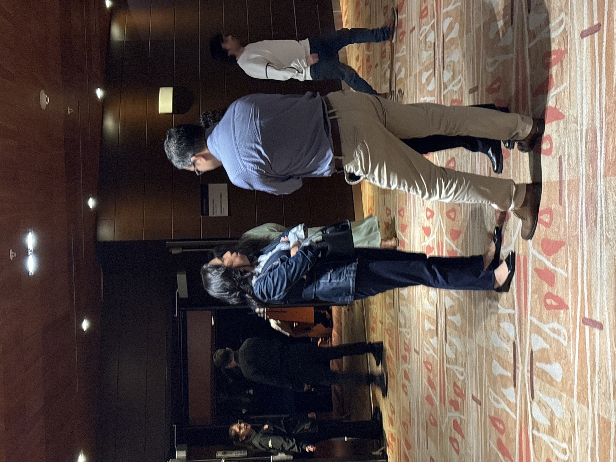 The image shows a group of people standing in an indoor space, likely a lobby or hallway.
A man wearing a light blue shirt and tan pants stands near the center of the frame, with his back to the viewer. To his right, partially overlapping, a woman in a denim jacket and dark pants stands beside a person in a green shirt. In the background, towards the left of the frame, two more individuals are partially visible.
Behind and to the left of the central figures is a dark brown paneled wall with sconce lighting. An informational sign is attached to the wall near the man in the light blue shirt. To the right of the central figures, the floor is covered with a patterned carpet featuring lines and abstract shapes in shades of beige, red, and white.
Based on the setting and the presence of what appears to be an informational sign, the location could potentially be a hotel, office building, or convention center in San Borja, Peru. However, the image lacks specific location details.