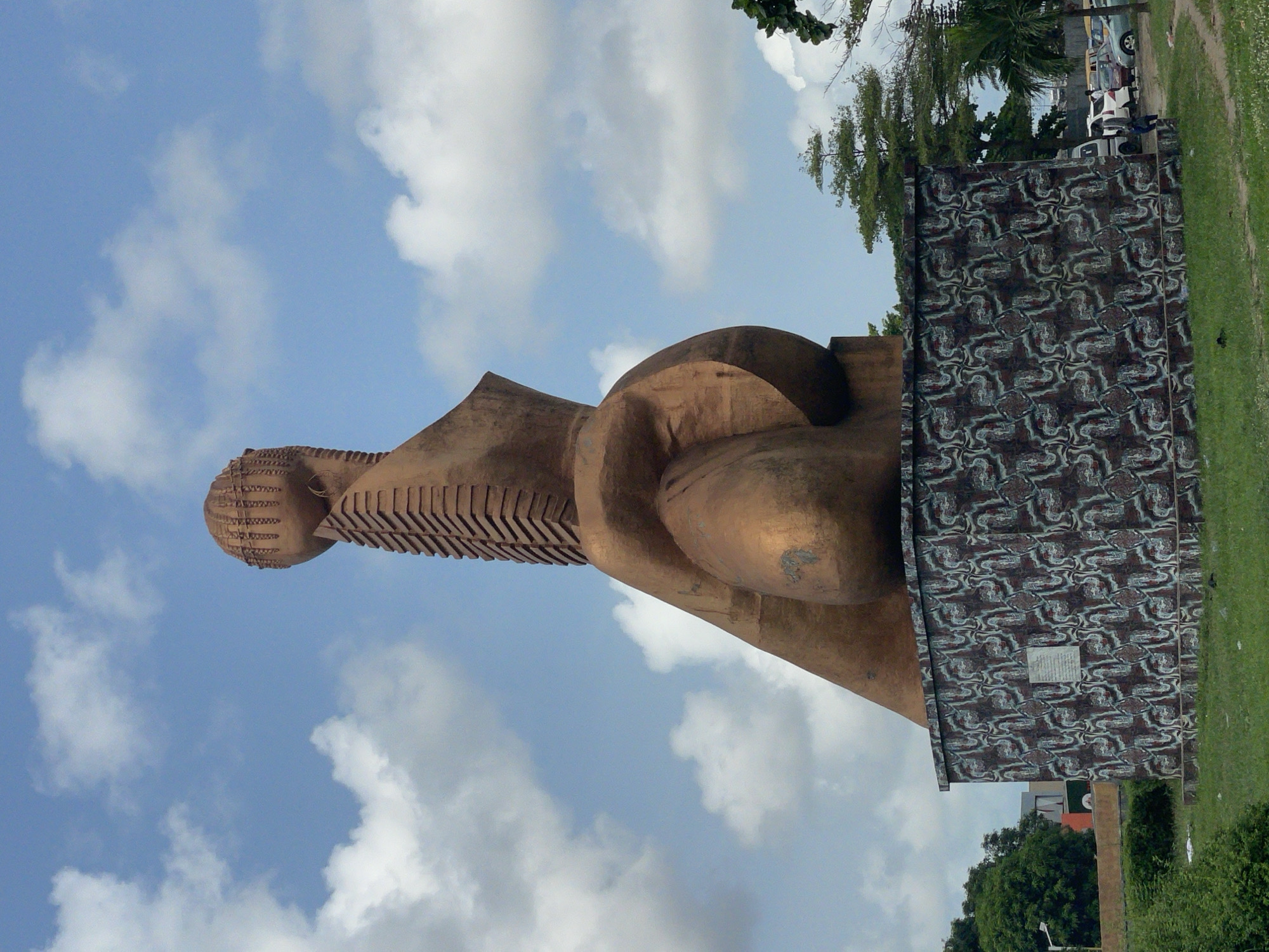 The image shows the Freedom Park monument in Lagos, Nigeria. It is a large, abstract bronze-colored statue with a patterned wall and base. The sky is blue with scattered white clouds. There is a small patch of green grass to the right of the monument. In the background, there are trees, cars, and other buildings.