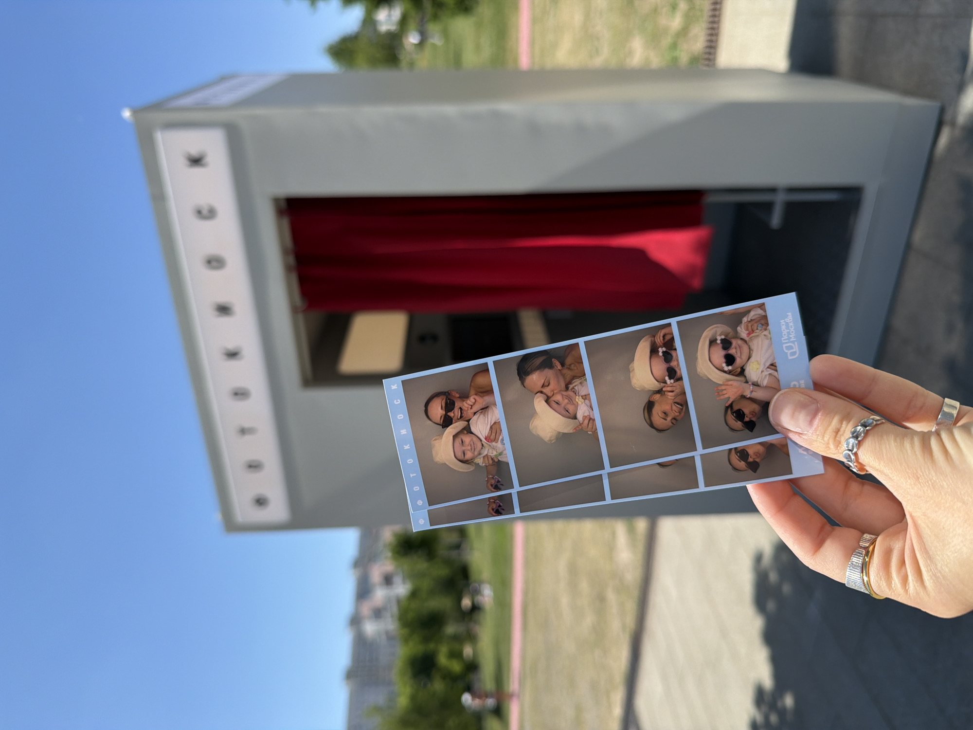 An outdoor scene captures a hand holding a photo strip in the foreground, set against a photobooth and a park landscape under a clear blue sky.
The hand, with fair skin, wears multiple rings: a wide, vertically grooved ring with a gold interior band on the index finger, a silver-toned textured ring and a plain silver-toned band on the middle finger, and a single silver-toned band on the ring finger. The hand holds a light blue-bordered photo strip featuring six individual photographs of a woman and a young child, likely a mother and daughter. In the photos, the woman, often wearing sunglasses, smiles and interacts affectionately with the child, who wears a light-colored bucket hat and heart-shaped sunglasses in some frames. The bottom left corner of the photo strip displays a logo and text reading "Парки Москвы," which translates to "Parks of Moscow."
Behind the hand and photo strip stands a grey, rectangular photobooth. Its top panel features black letters on a white background, spelling "ФОТОКИОСК" (PHOTOKIOSK). A red curtain is visible inside the booth's entrance.
The background depicts a sunny outdoor park setting. Green grass covers the ground, with trees scattered in the mid-distance. A pink-colored path runs horizontally across the mid-ground. Faint outlines of distant multi-story buildings are visible on a low hillside beyond the trees. The sky above is clear and bright blue, indicating good weather. Shadows suggest it is daytime. The location is identified as a park within Moscow, Russia, based on the text on the photo strip.