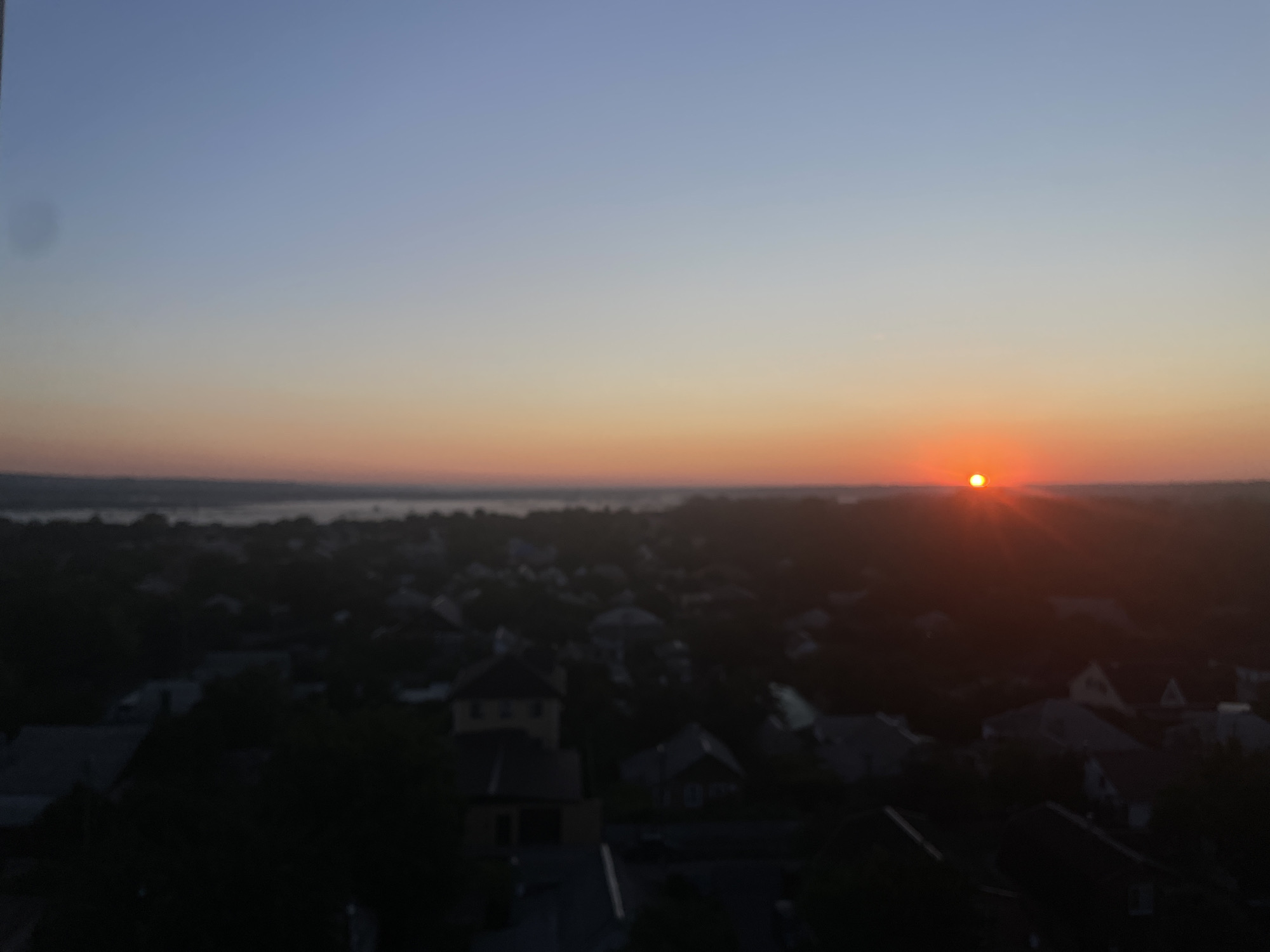 The image captures a sunrise over a residential area in Novocherkassk, Russia. The sun, a bright orange orb, is visible near the horizon, casting a warm glow across the sky which transitions from a pale blue at the top to a soft orange and pink hue closer to the horizon. The foreground is dominated by a dark silhouette of buildings, likely houses, suggesting this is a view from an elevated position, possibly a building or hill overlooking the town. The details of the buildings and landscape are obscured by the low light conditions and slight blurriness, but the general impression is one of a quiet, awakening urban or suburban environment. A body of water, possibly a river or lake, can be faintly seen in the distance on the left side of the image, further adding to the sense of a tranquil morning scene.