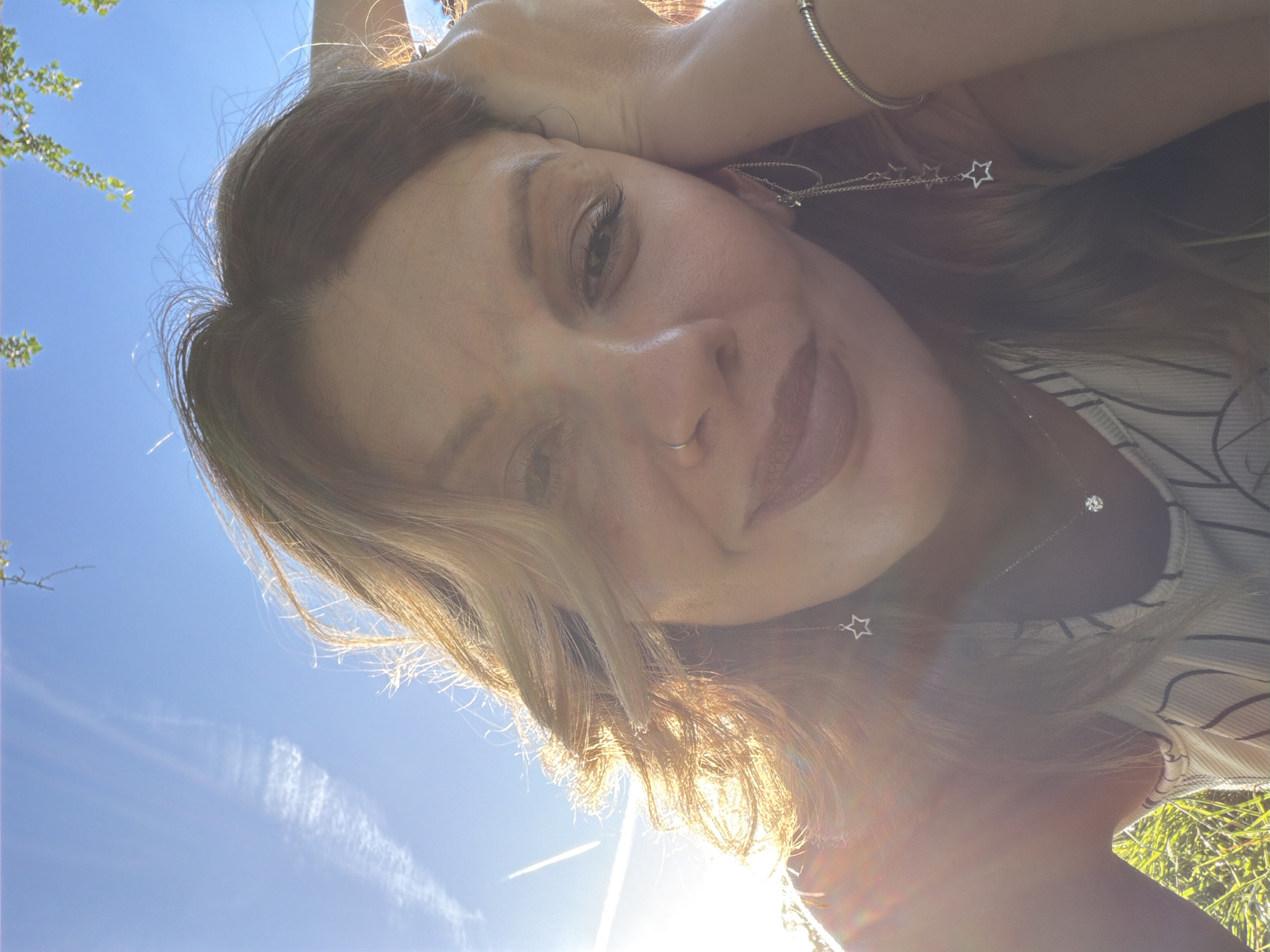A woman is seen in a close-up, low-angle shot, possibly a selfie, under bright sunlight. She has fair skin, light brown hair with blonde highlights, and is wearing a nose ring, a silver bracelet, and star-shaped dangling earrings. Her expression appears relaxed and smiling. She is wearing a striped top. The background includes a clear blue sky with faint contrails of a plane and some green foliage, suggesting an outdoor location, possibly a park or garden. No specific location in London, UK can be determined from the image.