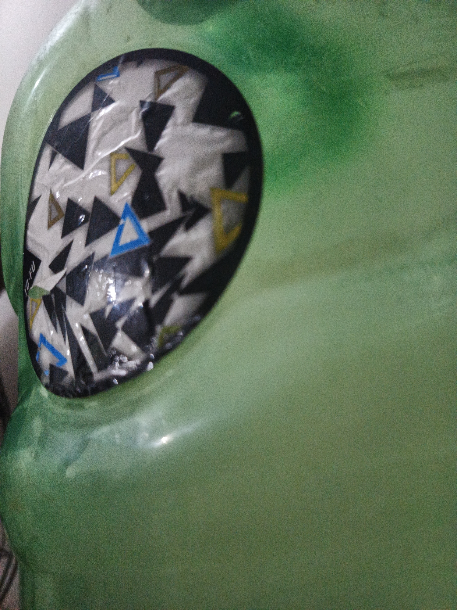 A close-up image shows a portion of a green, possibly plastic, object. A round sticker is affixed to the object. The sticker has a white background with black, yellow, and blue triangle patterns. The text "YO-FU" is visible on the left edge of the sticker. The scene's location in Los Robles, Venezuela is unknown. The content of this image suggests an indoor setting, focusing on the details of the green object and its sticker.