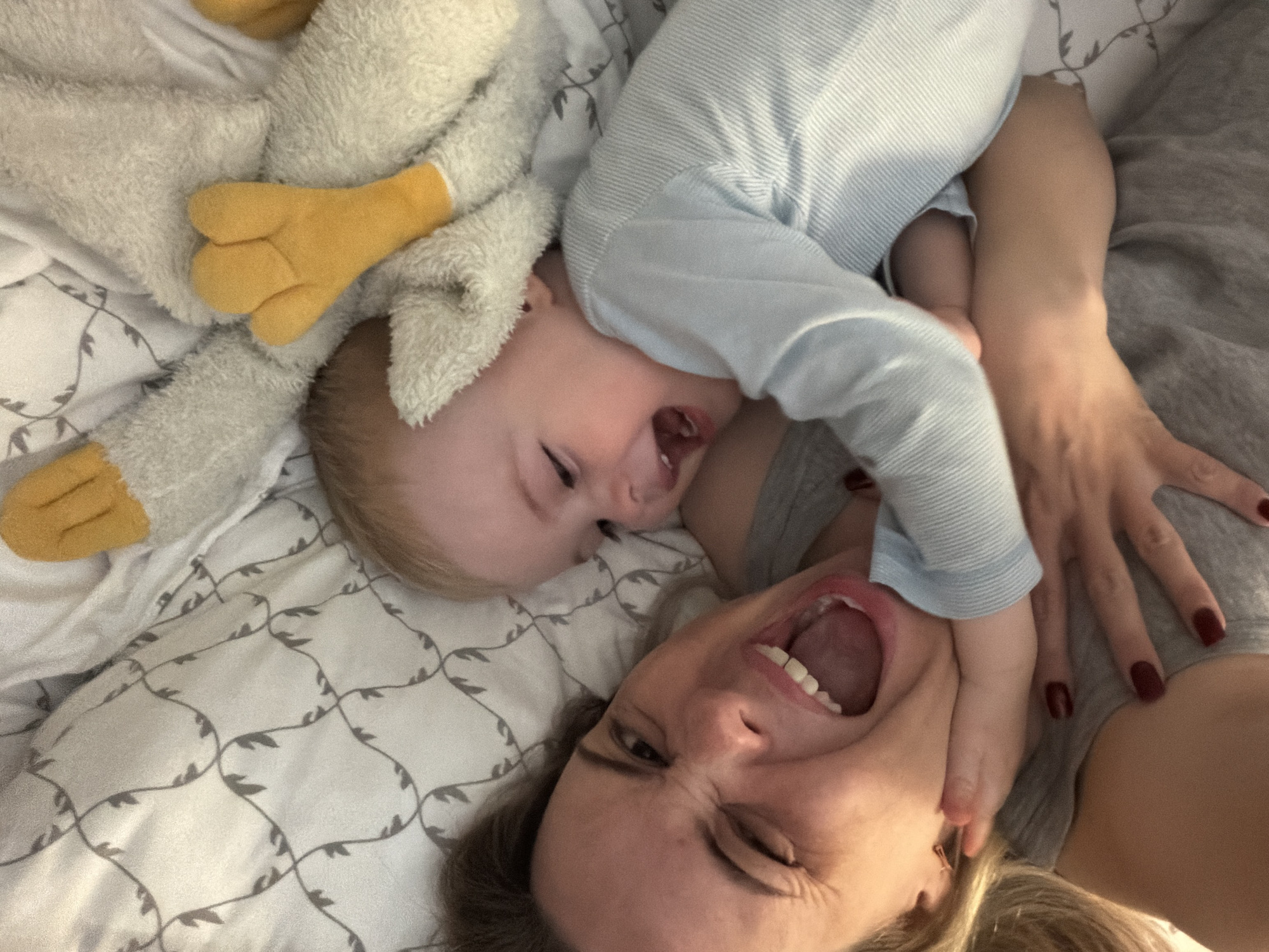 A woman and a baby are lying on a bed with a patterned white and gray sheet. The baby, wearing a light blue long-sleeved shirt, is positioned near the woman's face. The woman is lying with her head close to the bottom of the frame and is making an exaggerated smiling face. The baby is facing to the left and appears to be laughing as well. Beside the baby, there is a plush toy with yellow feet. The location is unknown and could not be verified by the image, nor could it be confirmed that the image was taken in Votkinsk, Russia.