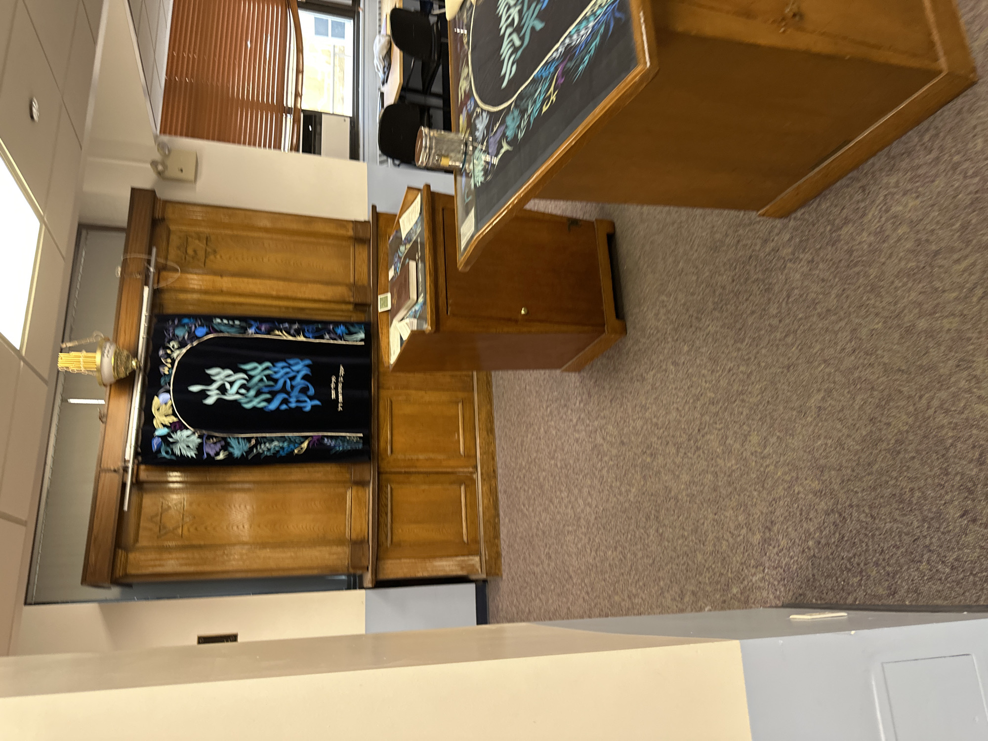 The image captures the interior of a Jewish religious or study space, likely a synagogue or prayer room. The room features a light brown speckled carpet, white walls, and a drop ceiling with recessed lighting.
Dominating the left side of the frame is a large, wooden Aron Kodesh (Holy Ark), distinguished by its carved panels. A rich, dark blue velvet parochet (Ark curtain) covers the central opening of the Ark. This parochet is elaborately embroidered with intricate floral designs and stylized Hebrew calligraphy in shades of light blue, green, and gold. The visible Hebrew script on the parochet appears to be a religious inscription. To the immediate left of the Ark, an ornate brass Ner Tamid (Eternal Light) is suspended.
In front of the Ark stands a wooden Bima or reading table, which is draped with a decorative cloth matching the parochet's design and featuring similar stylized Hebrew text. Several books, presumably prayer books or religious texts, are placed on this table. To the right of the Bima, another wooden cabinet or lectern is visible.
In the background, seen through an opening or doorway on the right, is an adjacent room furnished with chairs and tables, suggesting an office or study area. A window with light brown Venetian blinds is also partially visible in this background section. No individuals are present in the scene. The space is well-lit by interior lighting, making the time of day indeterminate. The overall ambiance is one of quiet solemnity, indicative of a place for religious observance or study.