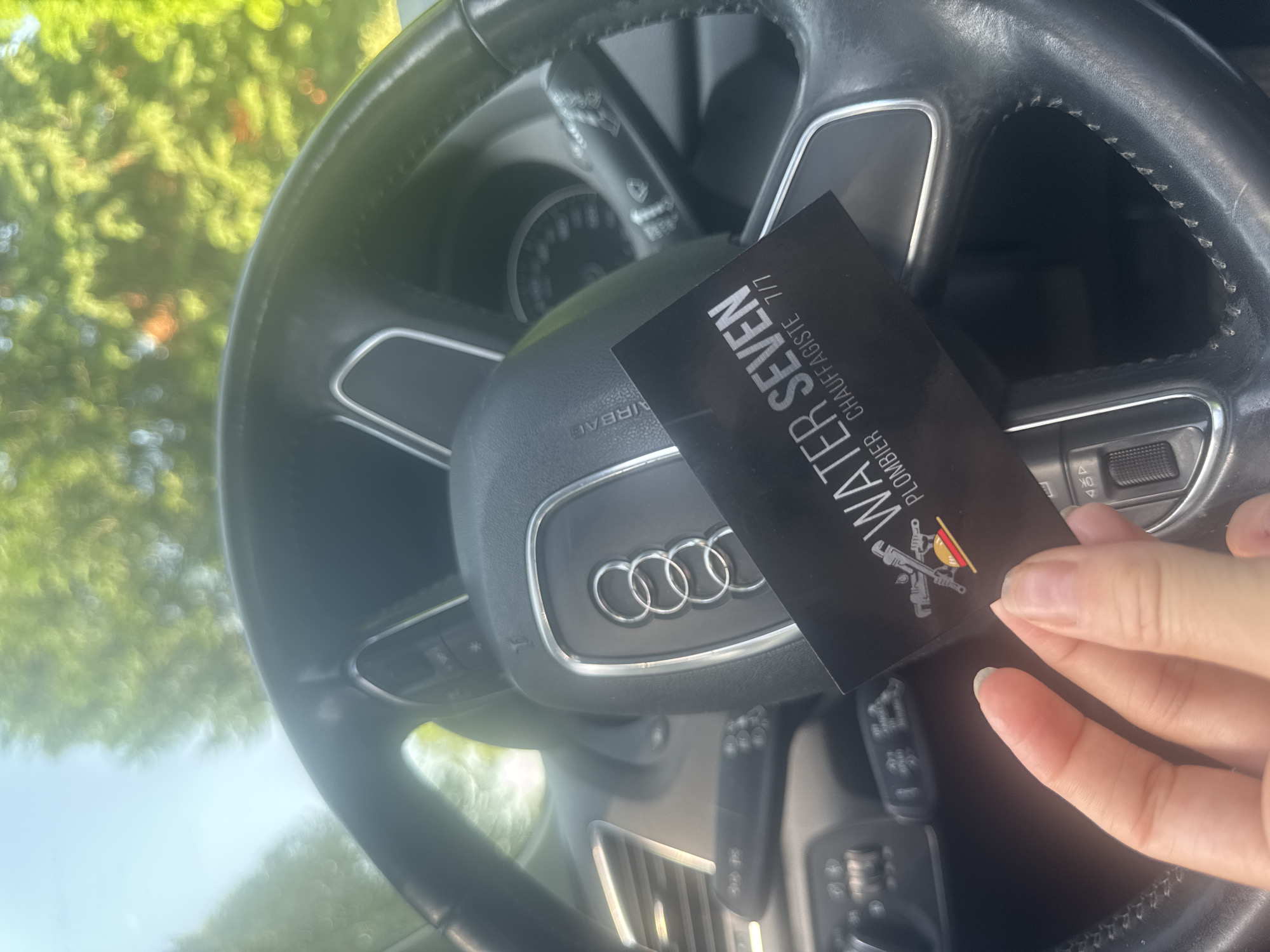 A hand holds a business card in front of an Audi steering wheel. The card reads "WATER SEVEN, PLOMBIER CHAUFFAGISTE 7/7" with a logo of plumbing tools. The environment is the interior of a car with a view of trees and daylight through the windshield. The city and country are not discernible from the image.