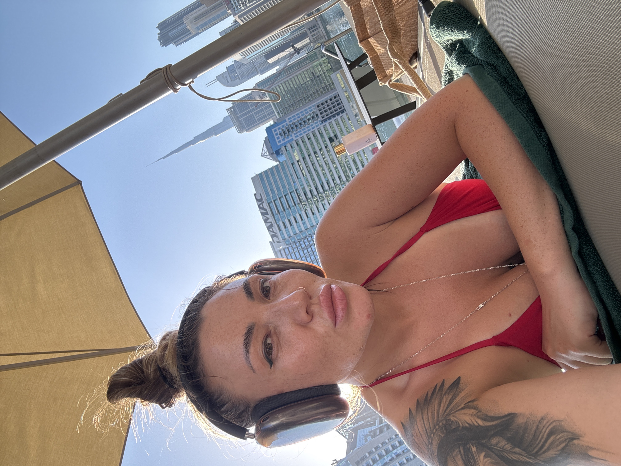 A woman is lying on a lounge chair, wearing a red bikini, headphones, and a silver necklace. Her hair is in a bun. She has a tattoo on her upper arm. The background shows a cityscape, specifically the Burj Khalifa and surrounding buildings in Dubai, United Arab Emirates. A portion of a beige sun umbrella and a green towel are also visible. The scene appears to be taken during daylight hours.