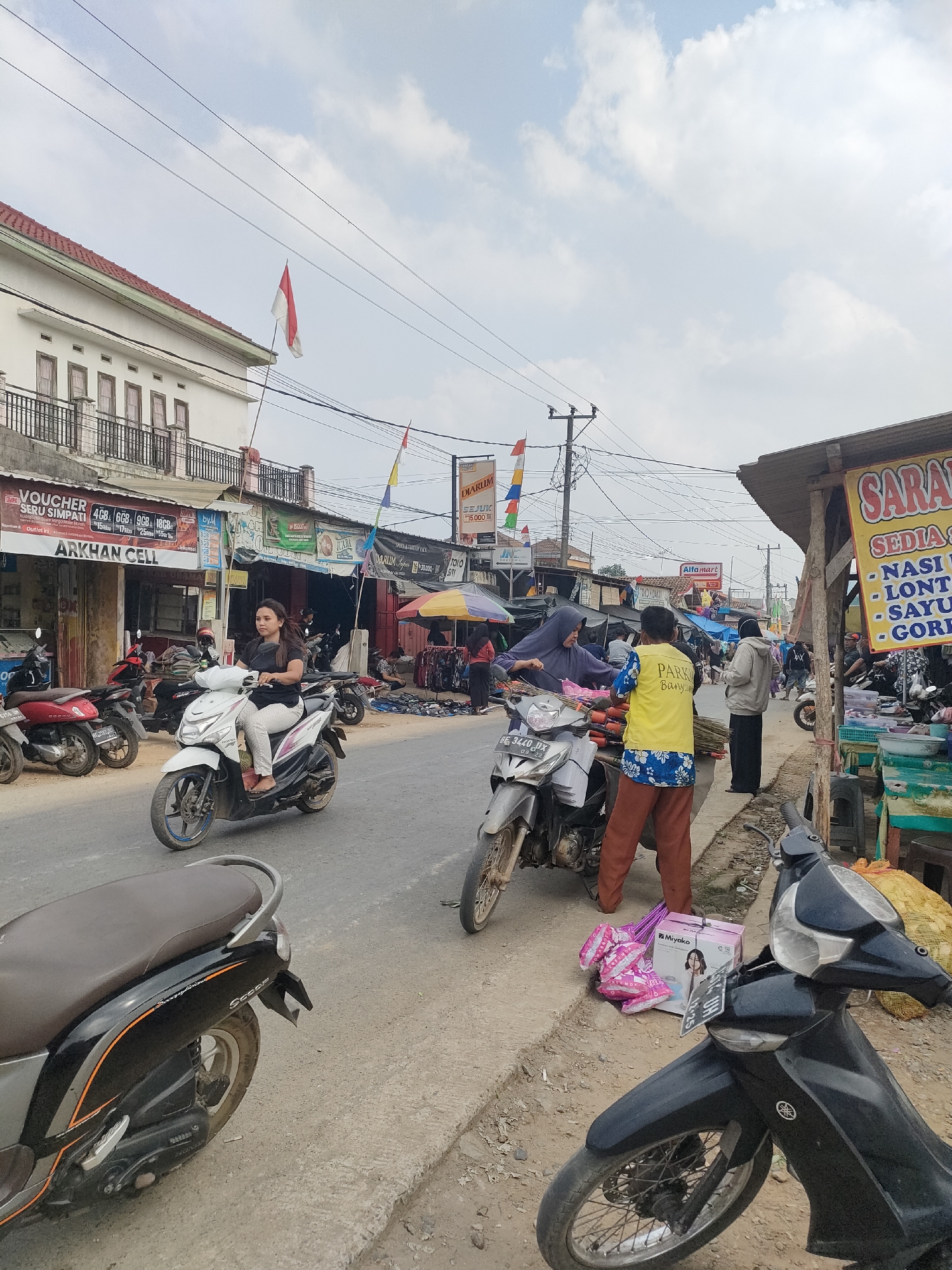 This is a street scene in Sukoyoso, Indonesia, under a partly cloudy sky. A paved road runs through the center, with a sidewalk to the right. On the left side of the road, there are buildings with shops. On the right side, there are market stalls.
Several motorcycles are parked and in motion on the road and sidewalk. A woman in a black shirt and patterned pants is riding a white scooter in the foreground, moving from left to right. Behind her, a silver scooter is parked on the side of the road. Further down the road, several other motorcycles are visible.
A group of people are gathered on the sidewalk to the right. One person, wearing a yellow vest with "PARKING" written on it, is interacting with another person in a purple hijab. A box labeled "Miyako" and pink balloons are on the ground near them.
Market stalls with various goods are set up along the sidewalk. One stall has a sign that reads "SARAI" and lists food items like "NASI," "LONT," "SAYU," and "GORENG." Another stall has clothing items displayed.
Indonesian flags are visible on poles above the shops on the left side of the street. Power lines crisscross the sky. The overall impression is a bustling commercial area with local vendors and daily life activities.