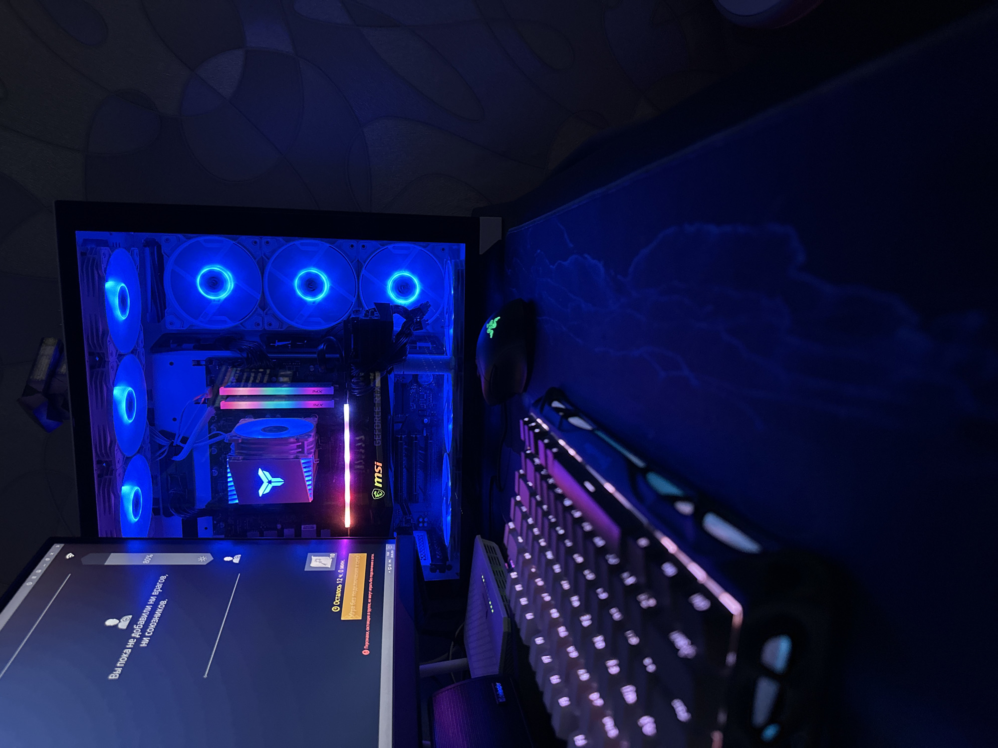 An overhead, medium shot captures a gaming setup in an indoor environment. The scene is dominated by the glow of RGB lighting emanating from the computer tower, keyboard, and monitor. The computer tower, positioned on the right, features illuminated blue fans and internal components. A keyboard with customizable RGB lighting occupies the foreground, with its keys displaying a purple and white glow. A dark computer mouse with green glowing accents is situated to the right of the keyboard. The monitor, on the left, displays a game interface with Russian text, indicating a game session in progress. The overall setting is dark, with the ambient light primarily coming from the electronic devices. The background appears to be a wall with a subtle textured pattern. The location is identified as Unknown, Unknown.