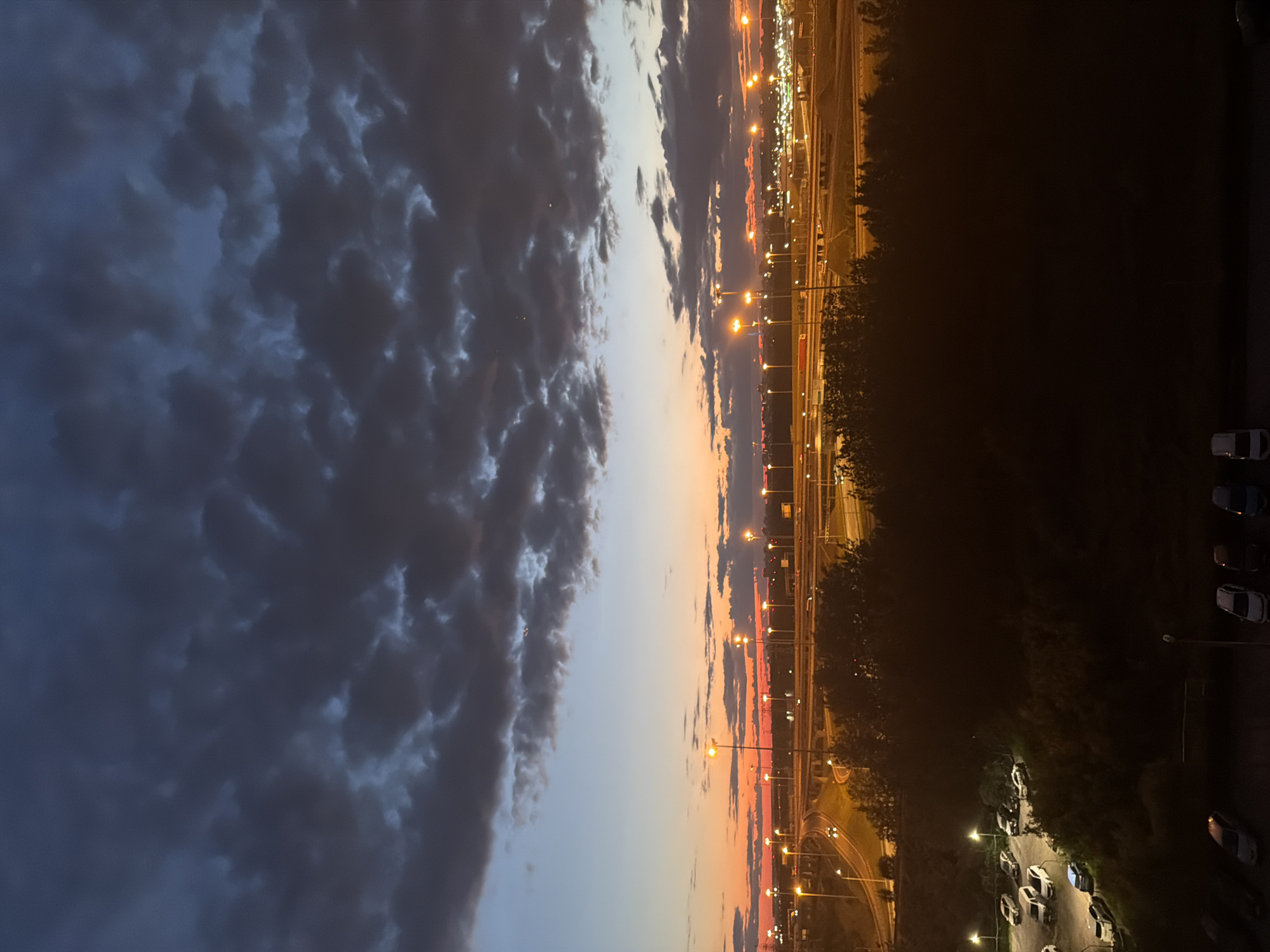 The image depicts a dusk scene in an urban environment. The sky is a gradient of dark blue clouds at the top, transitioning to lighter blue and then a vibrant orange and pink sunset near the horizon. In the mid-ground, a complex network of highways is visible, illuminated by streetlights and the headlights of passing cars, suggesting a bustling area. Beyond the highways, a cityscape stretches out, with more lights indicating buildings and urban infrastructure. In the foreground, a dark silhouette of trees or buildings obscures the lower part of the scene. A parking lot with several cars is visible to the left, illuminated by streetlights. The overall impression is of a city at twilight, with the transition from day to night captured in the sky and the artificial lights starting to dominate the landscape.
