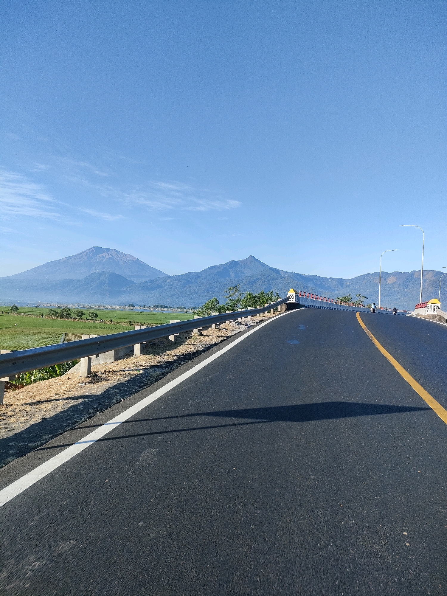 The image shows a road leading towards a mountain range under a clear blue sky. The road has a yellow dividing line and a white edge line. A metal guardrail runs along the left side of the road, separating it from a field of crops. In the background, two prominent mountains rise above a landscape of greenery and a body of water. The scene appears to be in Tambakboyo, Indonesia. Two motorcycles are visible on the road in the distance.