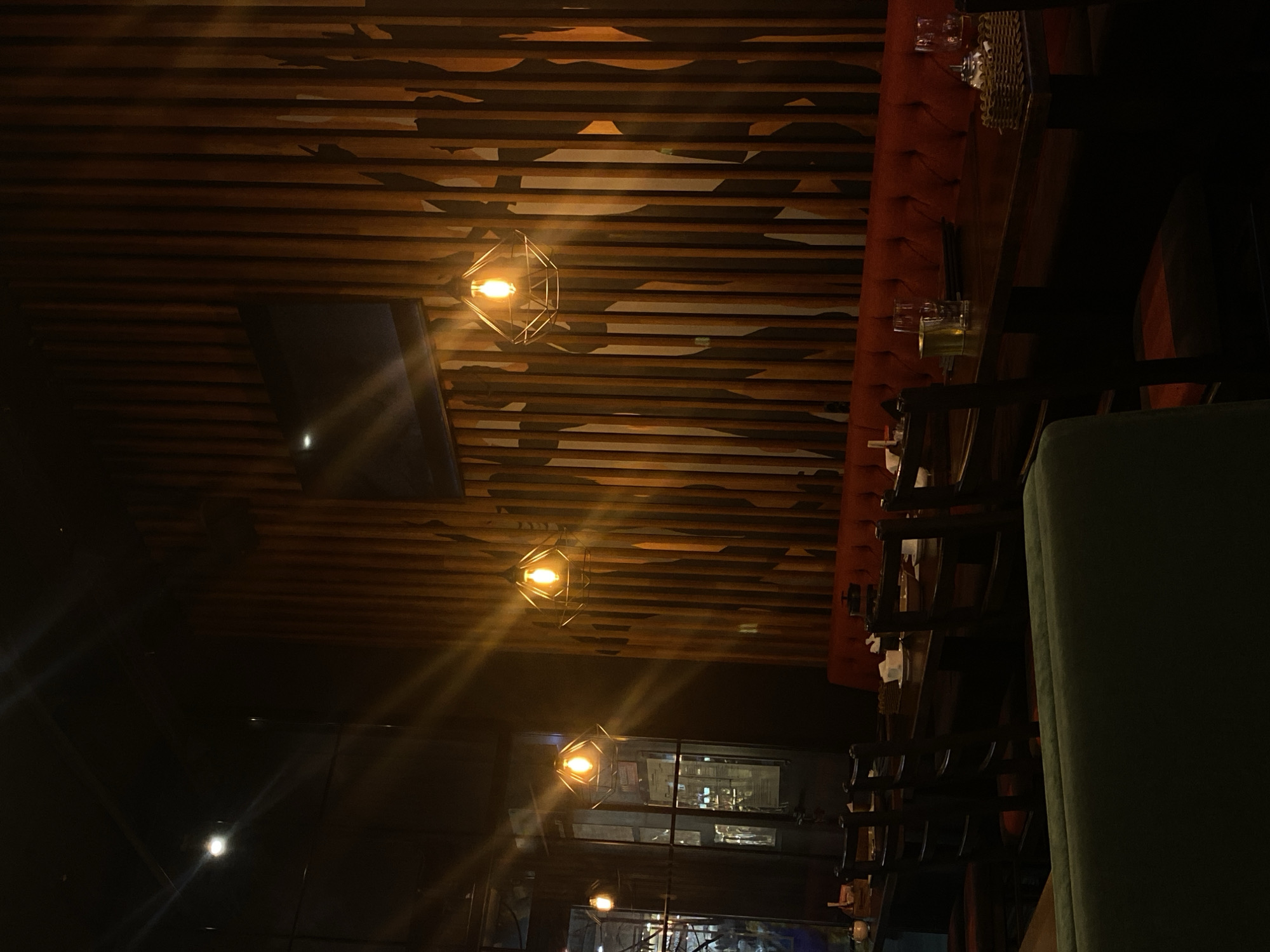 The image shows the interior of a restaurant or bar, possibly located in Kazan, Russia. The ceiling is decorated with horizontal wooden slats and three geometric, cage-like pendant lights. A dark screen is partially visible on the ceiling. A red upholstered bench runs along the right side, with wooden chairs and a table set with glasses and a woven placemat. The background shows a window to an exterior. The scene is dimly lit. A green chair is partially visible in the foreground.