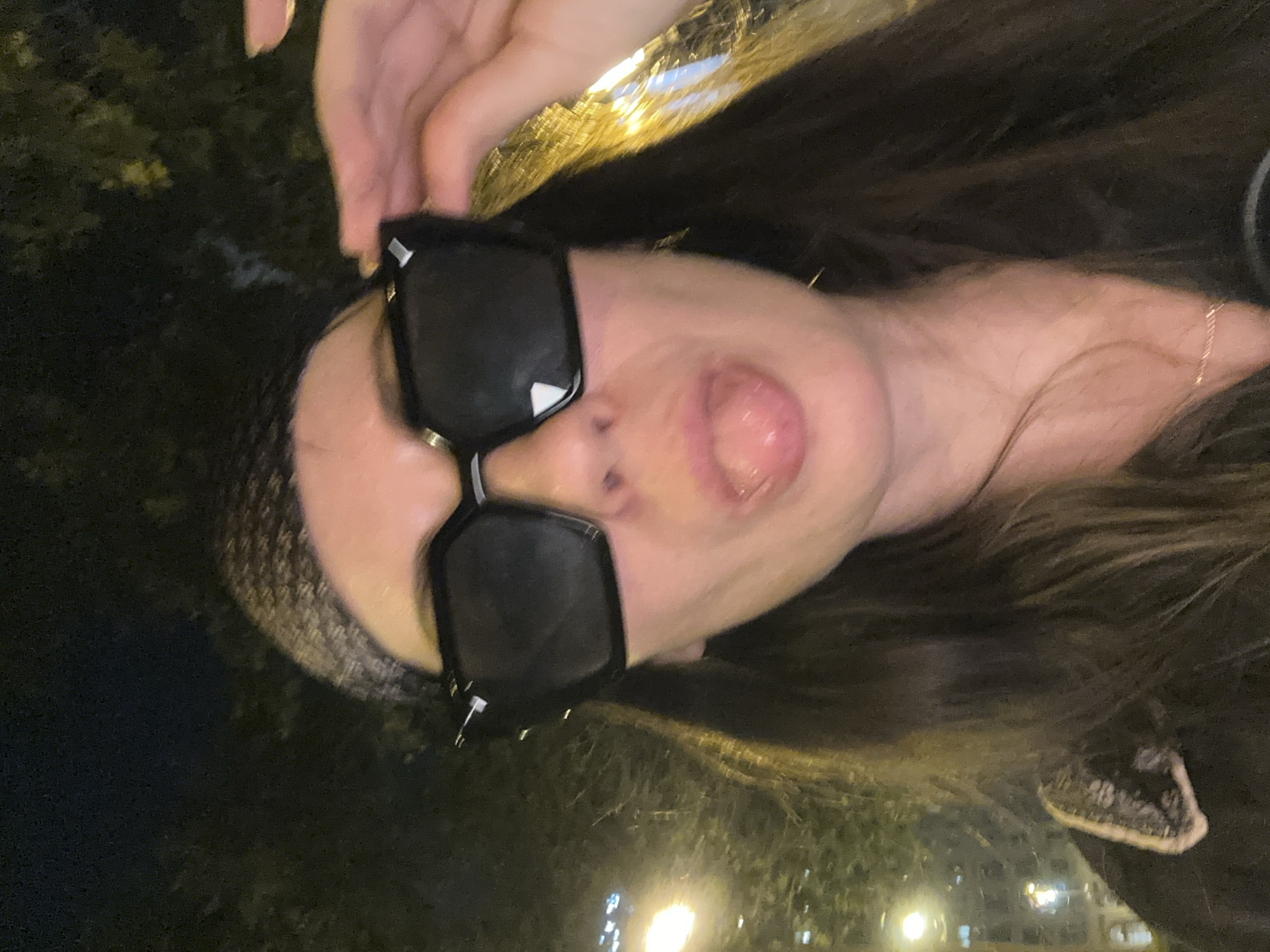 The image shows a close-up, sideways shot of a person wearing oversized black sunglasses. The person's mouth is open, with their tongue sticking out. They are wearing a dark head covering, and their long, dark hair is visible. A hand with manicured nails is touching the top of their head, near the sunglasses. The background is dark and blurred, with some faint lights visible, suggesting it is nighttime. The overall mood is playful and candid. The person appears to be outdoors, possibly in a city setting, with hints of vegetation and streetlights. Given the provided location context of Barcelona, Spain, the setting could be a street or outdoor venue in the city at night. There is no visible text in the image.