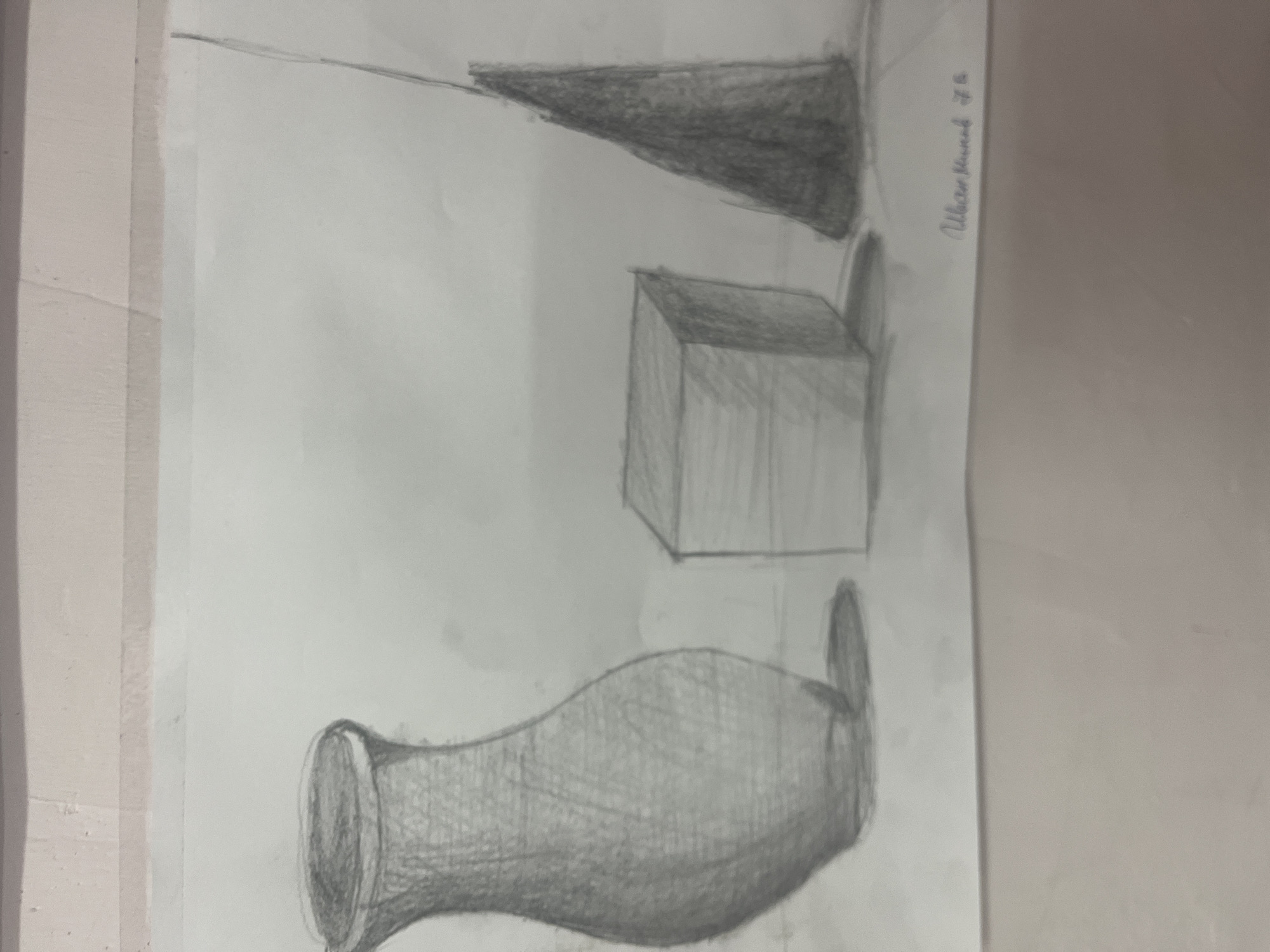 This media file features a pencil drawing of a still life composition, oriented horizontally on a sheet of light-colored paper. The drawing depicts three distinct objects, rendered with varying shades of gray to illustrate form and shadow.
In the bottom left, a vase or urn-like object with a flared top and a curved, bulbous body is shaded, suggesting a rounded surface and texture. It casts a shadow to its right. To the middle-right, a cube is drawn, showcasing three visible faces with differential shading that implies a light source from the upper left. This cube also casts a distinct shadow. Above the cube, towards the top right of the drawing, a cone is depicted lying on its side, with its base facing right and its tip pointing left. It too is shaded to give it volume and casts a shadow beneath it.
The overall scene of the drawing represents a still life study, likely an exercise in depicting three-dimensional objects and their interaction with light and shadow. The drawing technique involves pencil sketching and blending to create tonal values.
In the upper right corner of the drawing (when viewed horizontally), there is handwritten text in pencil, which appears to be a signature and a number, possibly "Ustaz Ahmed JJ 26" or a similar variant. The paper itself shows slight creasing and is situated on a light-toned surface. The location associated with this media file is Sofia, Bulgaria.