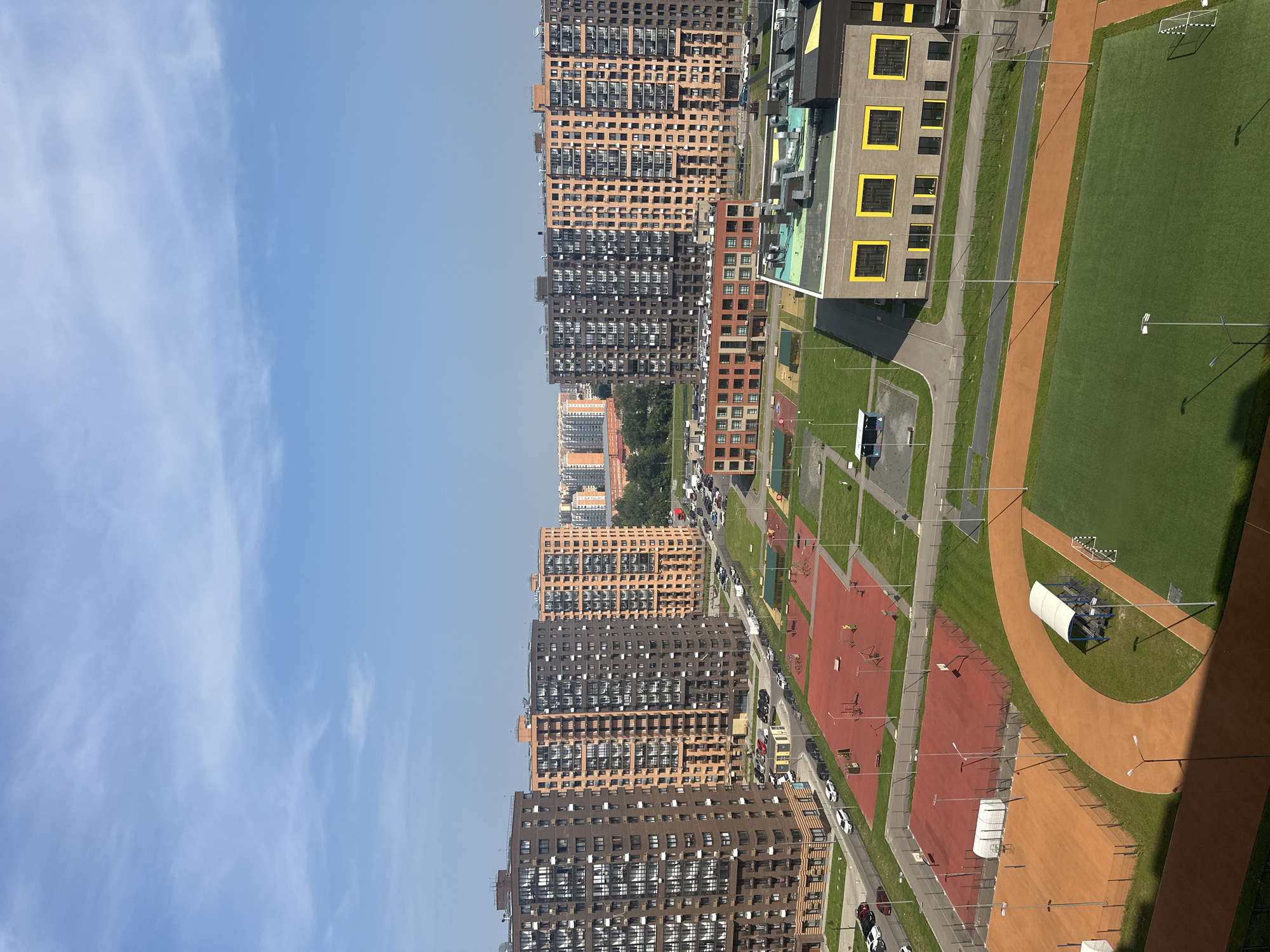 This aerial photograph captures a modern urban residential development in Balashikha, Russia, on a clear, sunny day with a few wispy clouds in the bright blue sky. The scene is dominated by tall, multi-story apartment buildings constructed with a mix of orange and dark grey brick facades and numerous balconies. In the foreground, a sports complex is visible, featuring a green soccer field with a goal, an orange running track, and a fenced-off tennis court. Adjacent to these are red-surfaced courts, possibly for basketball or volleyball, with various sports equipment like basketball hoops and playground structures. A low-rise, modern brick building with bright yellow window frames, likely a school or community center, is situated in the mid-ground. The streets are lined with parked cars, and trees are sparsely scattered throughout the area. The overall impression is one of a newly developed, organized, and functional urban environment designed for residential living and recreation.