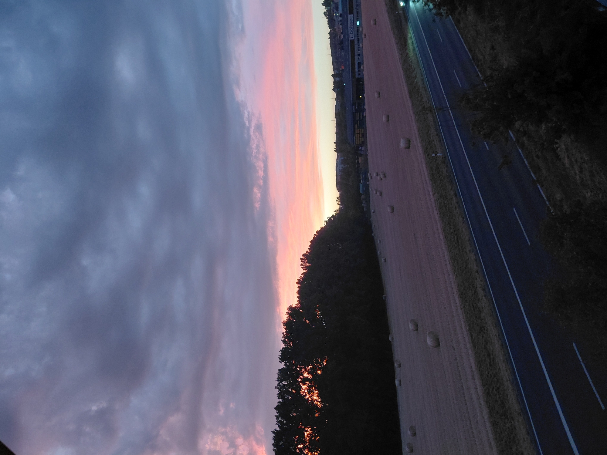 This is an image of a rural landscape at dusk. The sky is a gradient of colors, with dark gray clouds overhead transitioning to shades of pink and orange near the horizon. In the foreground, a mown field is scattered with round bales of hay. A dark road with white lane markings runs diagonally across the lower right portion of the image. Along the left side of the field, a dense line of trees silhouetted against the sky separates the field from a distant town. The town appears to have low-rise buildings, including one with a prominent sign that reads "Boissec". The scene is serene and peaceful, indicative of a rural setting.