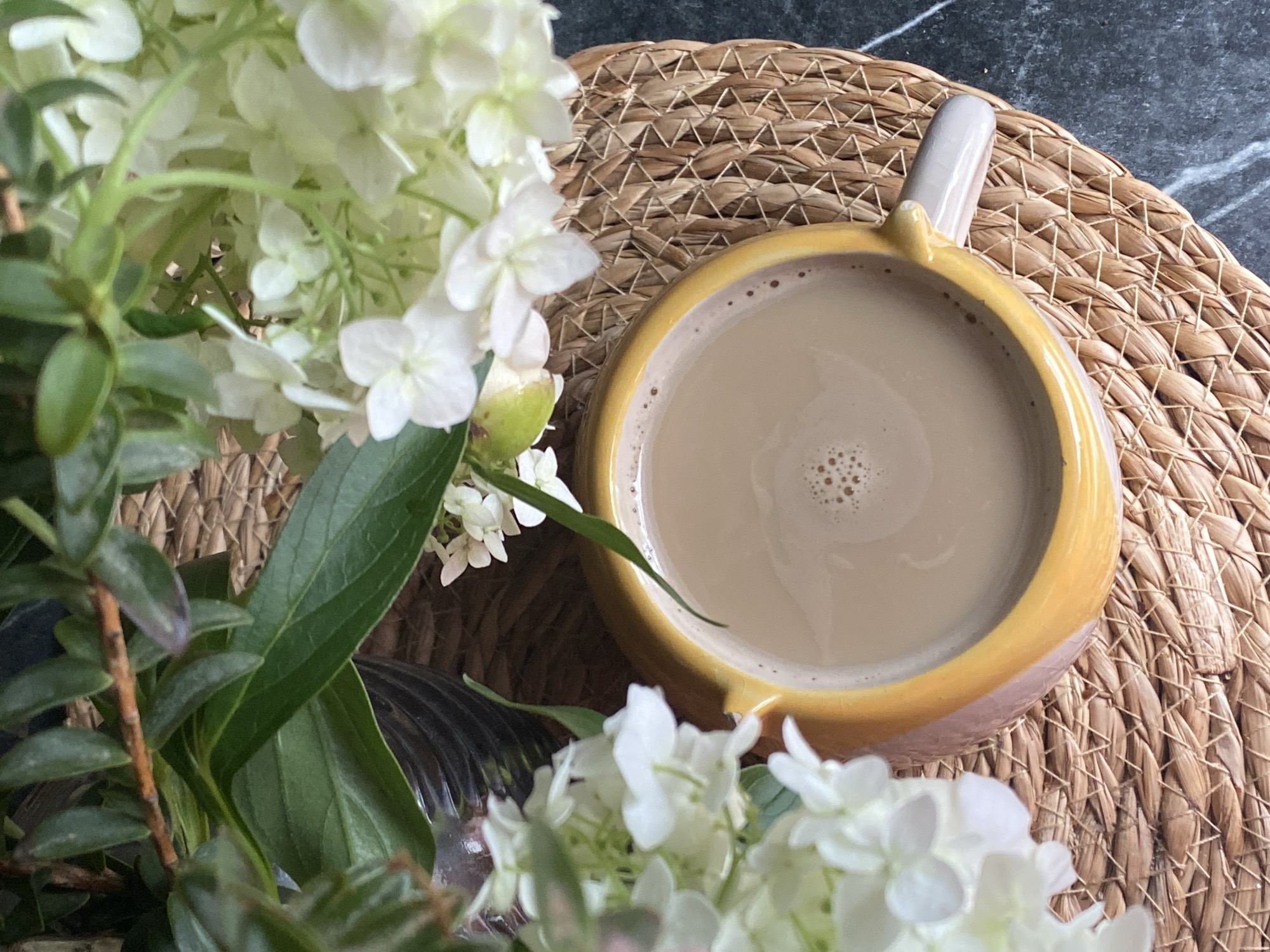 A close-up overhead shot shows a yellow ceramic mug filled with a light brown beverage, possibly coffee or tea, with a frothy surface. The mug rests on a woven circular placemat, which is situated on a dark, marble-like surface. To the left and top of the mug, a bouquet of white flowers and green foliage is visible. The scene appears to be an indoor setting, likely a home or cafe, with natural lighting. There are no people or animals present in the image. No specific location in Chaikovsky, Russia, can be identified from the visual information provided.