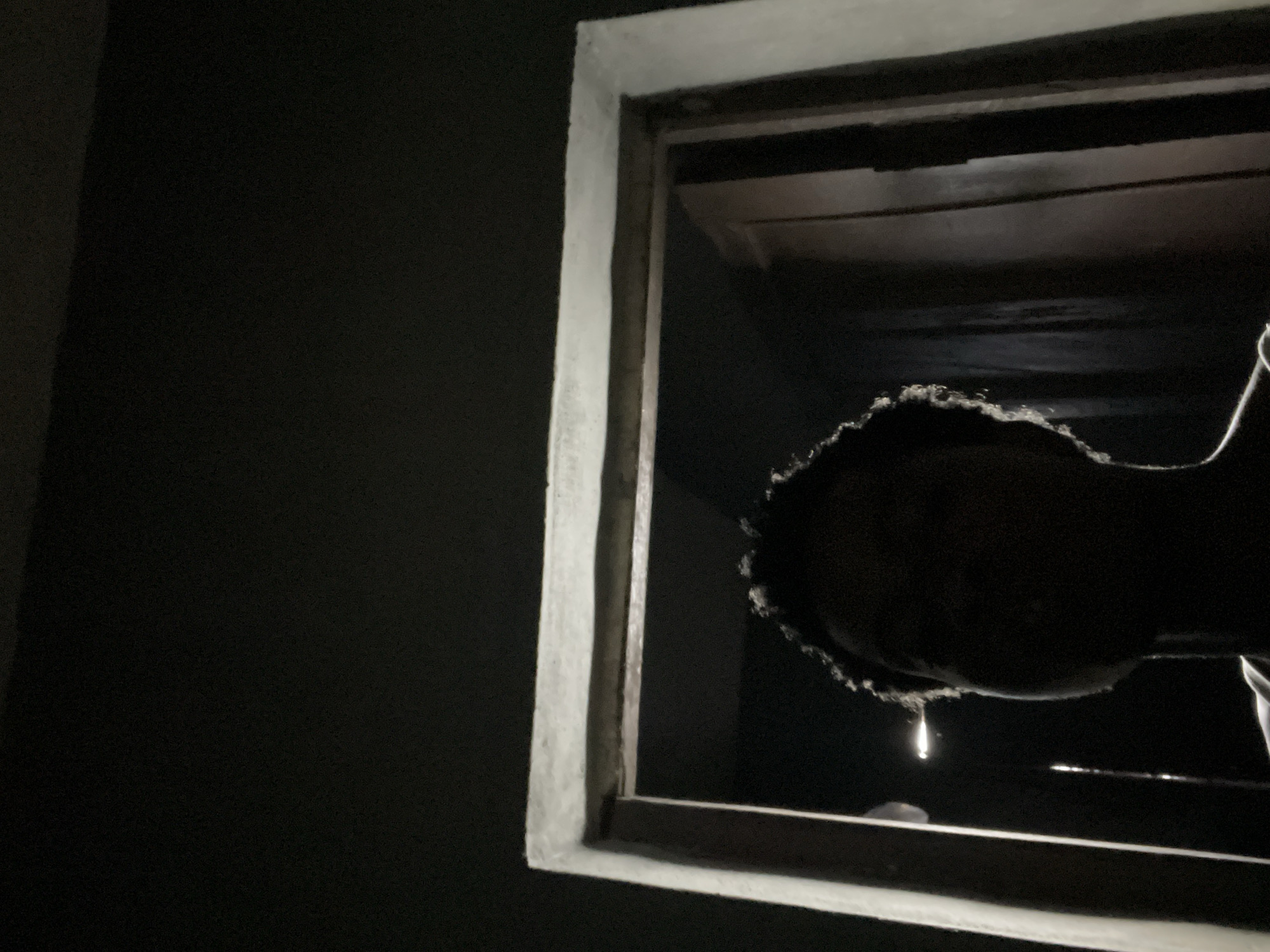 An individual's face, silhouetted against a dimly lit doorway in Nkpor, Nigeria, is the central focus of the image. The lighting suggests an indoor setting, possibly a home, with a strong light source emanating from behind the doorway, casting the individual's features into shadow. The texture of the individual's hair, with its defined curls, is slightly visible due to the backlighting, creating a halo effect around their head. The doorway itself appears to be wooden, with a white frame that is slightly worn. The overall impression is one of mystery and introspection, with the darkness obscuring details and leaving the viewer to infer the circumstances.