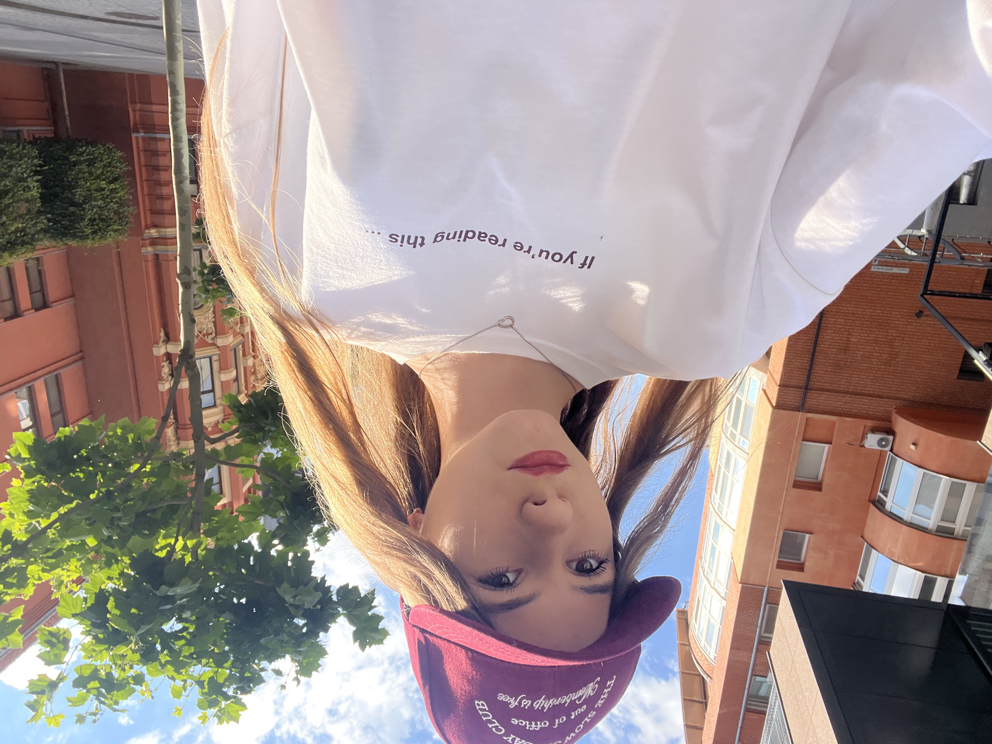 A young woman with long, light brown hair is taking a selfie outdoors. She is wearing a white long-sleeved shirt with text printed on it, and a maroon baseball cap. The shirt has a small, silver heart-shaped pendant necklace on it. The text on the shirt reads, "If you're reading this...". The text on the cap is partially visible and appears to read, "THE SLOW DAY CLUB", and "out of office membership is free".
The woman is looking directly at the camera with a neutral expression. Her makeup includes red lipstick and mascara.
The background shows buildings with red brick facades and white-framed windows. There are also trees with green leaves and a bright blue sky with some clouds, suggesting it is daytime and the weather is pleasant. The scene appears to be in an urban environment. The image is oriented with the top of the woman's head at the bottom of the frame.
The location context provided is Dnipro, Ukraine.