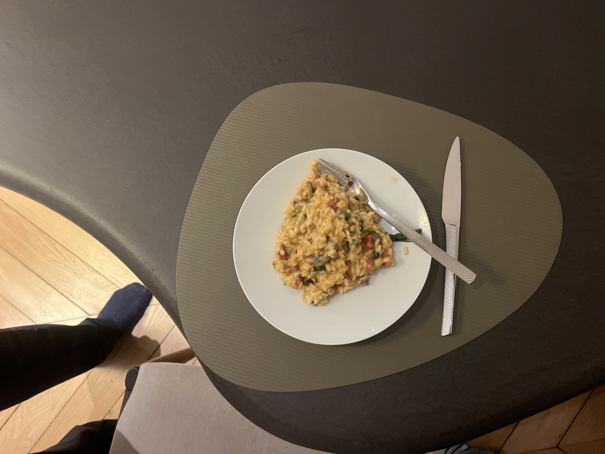 A top-down shot shows a plate of risotto with a fork and knife on a placemat on a dark surface. A person's legs are visible in the lower left corner, wearing a dark sock. The environment appears to be an indoor dining area, evidenced by the wooden floor and furniture. Without more information, the location in Paris, France cannot be determined.