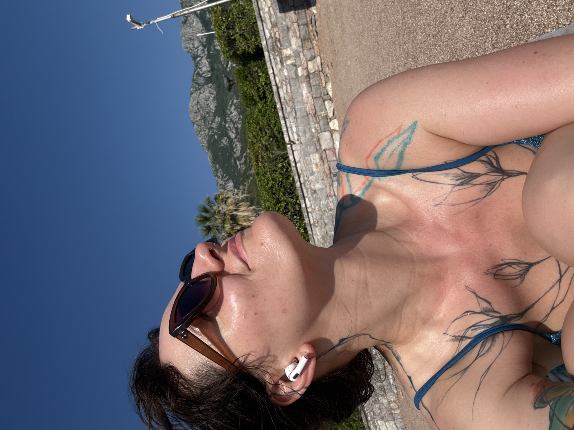 A woman with short, dark hair and sunglasses is shown in a close-up, low-angle shot, looking up towards a clear blue sky. She is wearing a blue bikini top and has several tattoos visible on her chest, neck, and shoulder. White earbuds are in her ears. The background consists of a beach, a stone wall, lush green bushes, palm trees, and rugged mountains under a bright sky. A lamppost is visible on the right side of the frame. The scene appears to be a sunny day in Sveti Stefan, Montenegro.