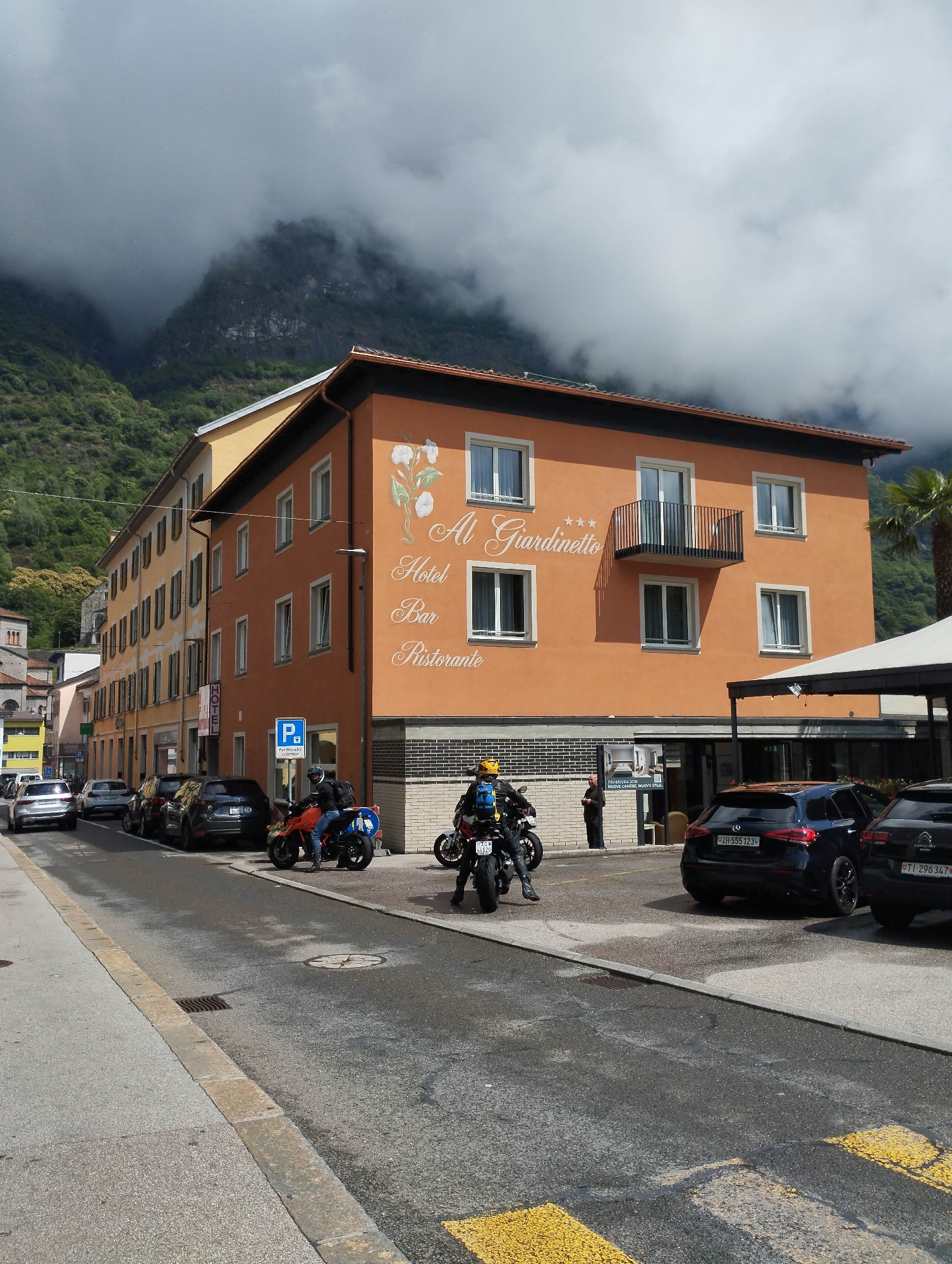 This detailed image depicts a street scene in a mountainous region, likely a town or village given the density of buildings. The sky is overcast with thick, low-hanging clouds and fog partially obscuring the upper portions of steep, green-forested mountains in the background. The central feature is a three-story building with an orange-ochre facade, distinguished by white decorative script that reads "*** Al Giardinetto Hotel Bar Ristorante," accompanied by a painted motif of white flowers and green leaves. The building features multiple white-framed windows, with one on the second floor having a small black wrought-iron balcony. Its roof has reddish-brown tiles and dark trim. Below the "Ristorante" text, a sign advertises "PRIMAVERA 2020 NUOVE CAMERE, NUOVO STILE" (Spring 2020 New Rooms, New Style). Adjacent to this, on the left, is a lighter yellow-ochre building of similar height, displaying a vertical "HOTEL" sign and numerous windows with dark, closed shutters. Further down the street on the left, additional buildings of varying colors (yellow, light pink, white) are visible, with a distant hint of a church tower. In the mid-ground, a paved road stretches towards the background. Several vehicles are parked along both sides of the street. On the left, a silver SUV and a dark grey hatchback are visible, followed by a dark SUV (possibly a Mazda CX-5). On the right, in a designated parking area in front of the orange hotel, two motorcycles are prominent. An orange KTM-style sport bike is being ridden by a person wearing a black jacket, blue jeans, a yellow helmet, and a blue backpack, with their feet on the ground. A blue parking sign indicating "Particolare clientela" (specific clientele) is visible near this motorcycle. Another person, dressed in dark attire, stands near the hotel entrance. A dark blue Mercedes-Benz A-Class (Swiss license plate: ZH-555 123 from Zurich canton) and a dark Citroen C3 Aircross (Swiss license plate: TI-296 347 from Ticino canton) are parked behind the motorcycles. A large white canopy structure extends from the orange building over part of the parking area on the right. The immediate foreground features a concrete sidewalk with a raised, rough-hewn curbstone of yellowish-brown stone. The asphalt road surface shows some dark, wet-looking patches and a storm drain grate. Yellow painted lines, possibly indicating a crosswalk, are visible at the bottom right. A tall palm tree is partially visible behind the orange building on the far right. Overhead wires