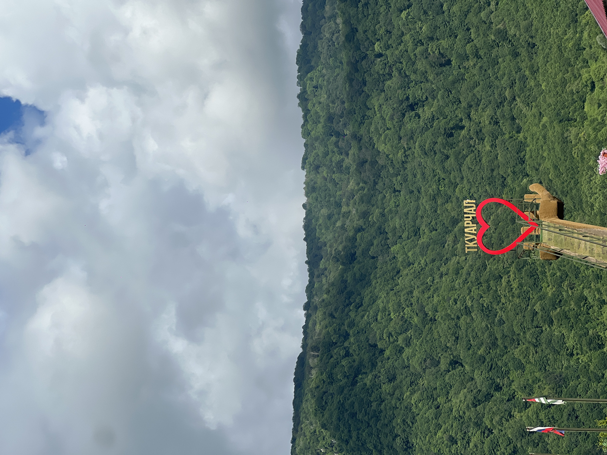 A large, red, heart-shaped sculpture with the word "ТКУАРЧАЛ" in yellow lettering above it is situated on a mountainous slope covered in dense green trees under a cloudy sky. The sculpture appears to be a tourist attraction. In the foreground, two flags are visible on poles to the left of the sculpture, and a section of a walkway or platform leads up to the heart. The overall impression is that of a scenic, possibly rural, location.