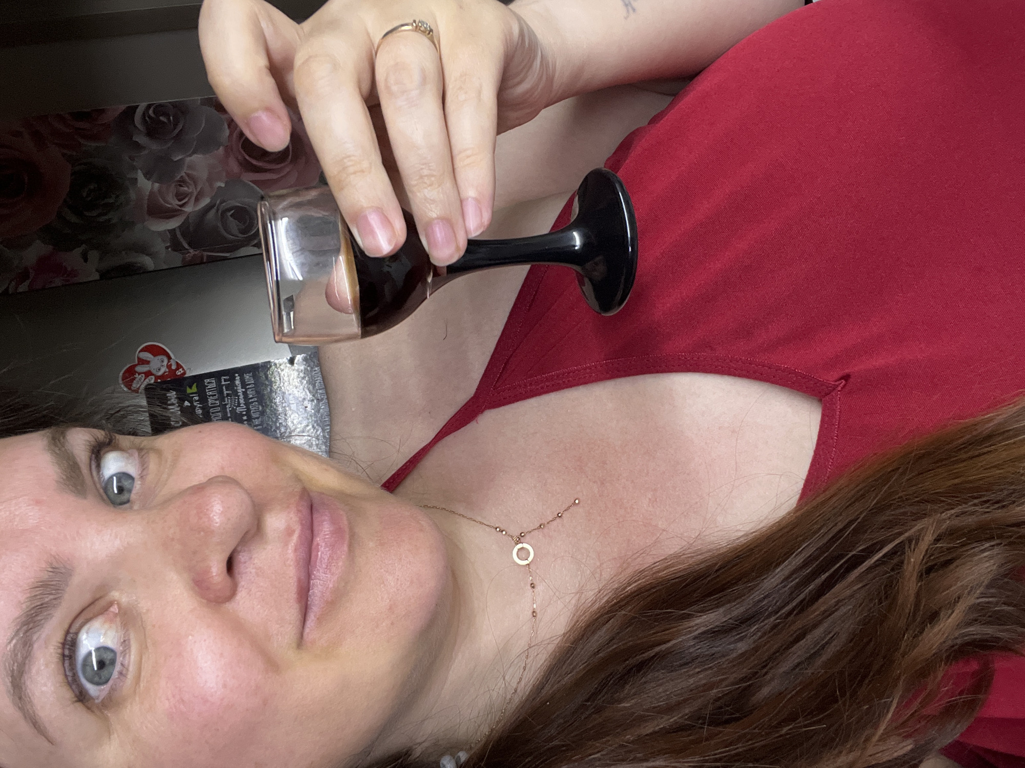 The image shows a close-up of a woman with light skin, brown hair, and blue eyes. She is wearing a red tank top and a delicate gold necklace with a circle pendant. The woman is holding a small shot glass filled with a dark liquid, possibly espresso or liqueur, in her right hand. Her gaze is directed upwards and to her left, with a slight smile. In the background, there is a refrigerator with a floral-patterned magnetic frame and some stickers. The overall setting appears to be a kitchen in a home environment. There is no indication of the specific location within the city of Noviy, Russia.