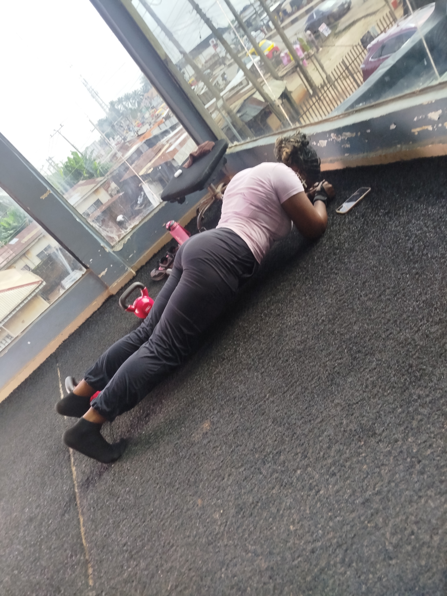 In the city of Uselu, Nigeria, an individual is engaged in a plank exercise on a gym floor situated near a large window. The person is wearing dark grey sweatpants and a light pink t-shirt, with black socks on their feet. Their body is positioned horizontally, parallel to the floor, supported by their forearms and toes. Next to the individual, on the floor, are a red kettlebell and a pink water bottle. A smartphone is also visible on the floor. Through the window, a street scene with buildings, vehicles, and overhead electrical wires is visible. The lighting suggests it is daytime.