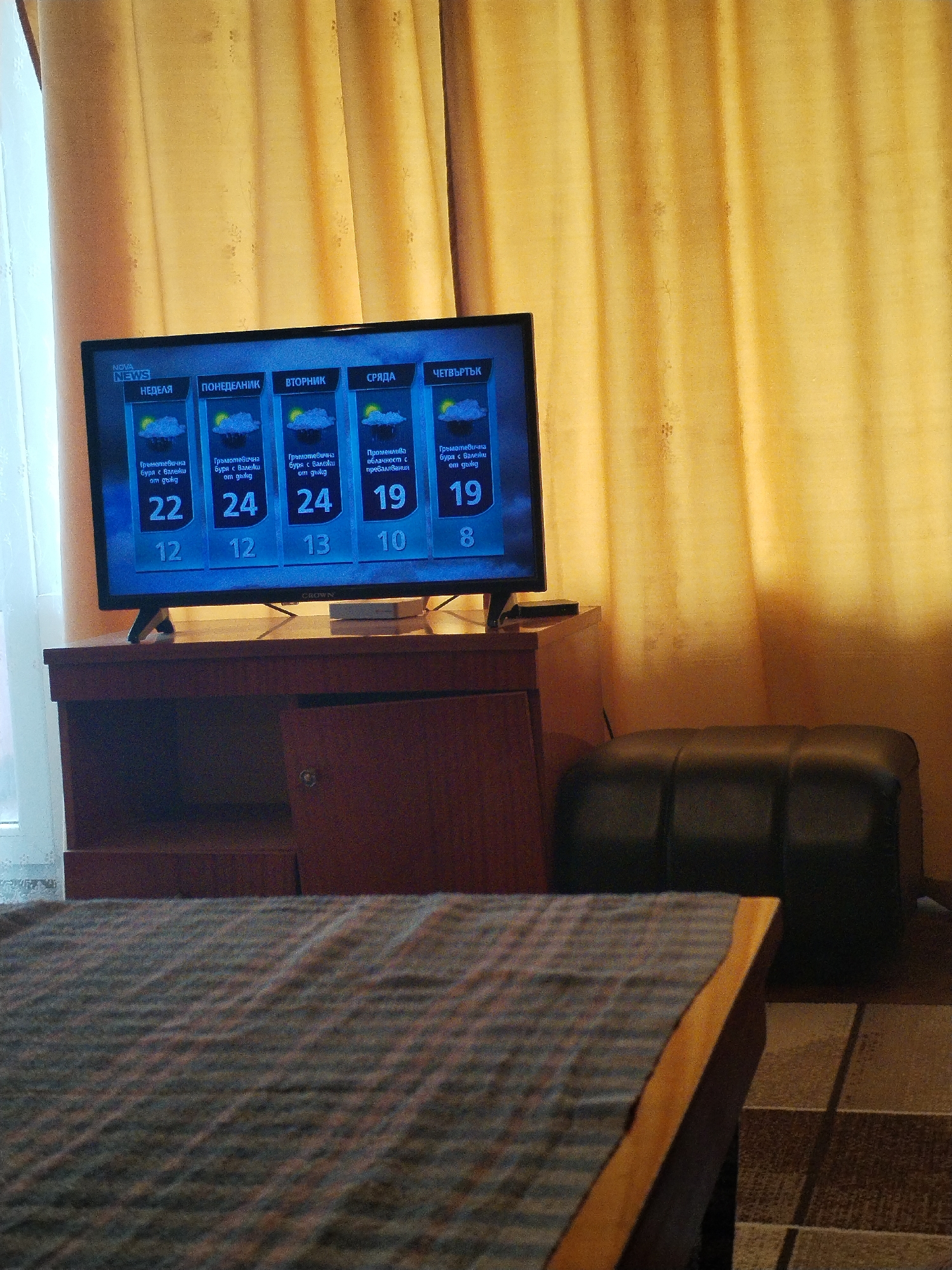 The image depicts an indoor scene, likely a hotel room in Varna, Bulgaria. The foreground shows a blurred table corner, leading to a dark-colored ottoman. Behind the ottoman is a wooden TV stand. Atop the stand is a flat-screen TV, displaying weather forecast in Bulgarian. The forecast predicts thunderstorms and showers throughout the week. The background features golden/tan colored curtains, slightly obscuring a window with faint light coming through.