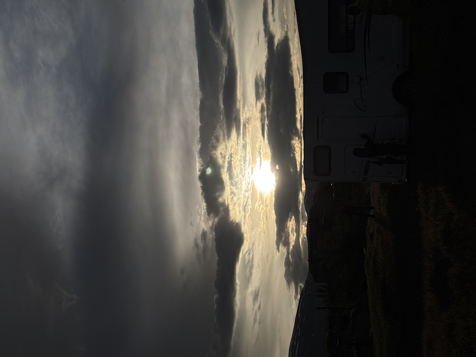 The image shows a dramatic sky with the sun visible through breaks in dark, striated clouds. A recreational vehicle (RV), with a partial view of the word "Classique" on its side, occupies the right side of the frame. Silhouetted figures of two individuals appear near the RV, and an area of dark, possibly grassy or overgrown ground is visible in the foreground. The location, Anipemza, Armenia, is not verifiable from the image content.
